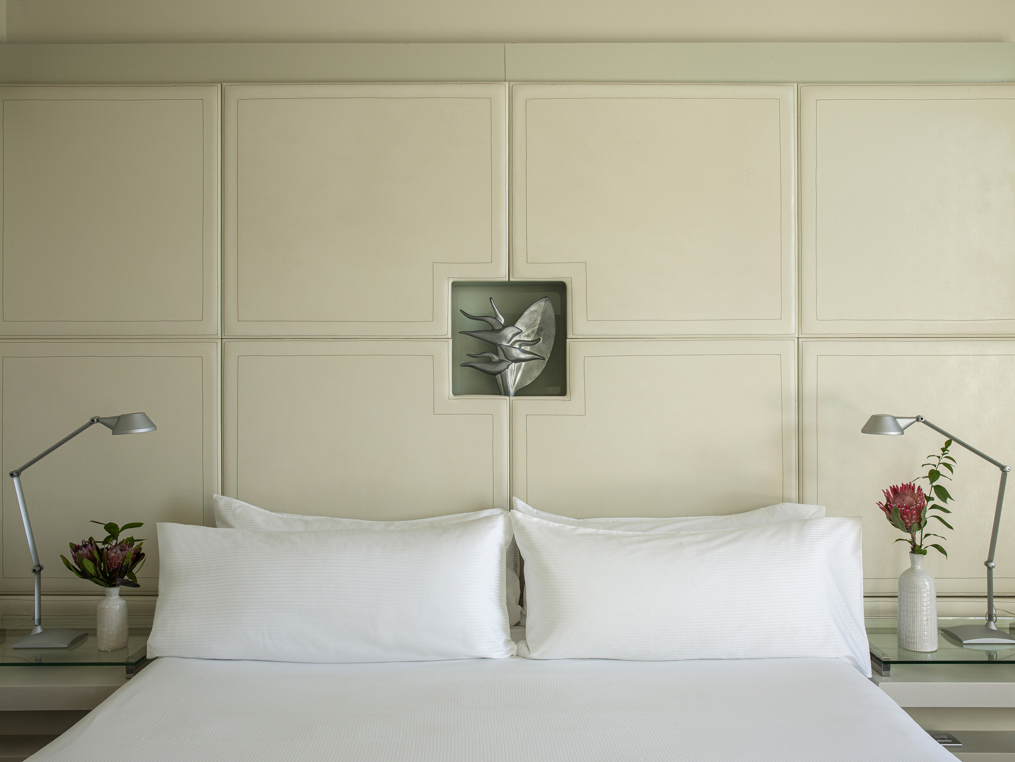 Hotel bedroom with white bed, paneled headboard, metallic art, and protea flowers.