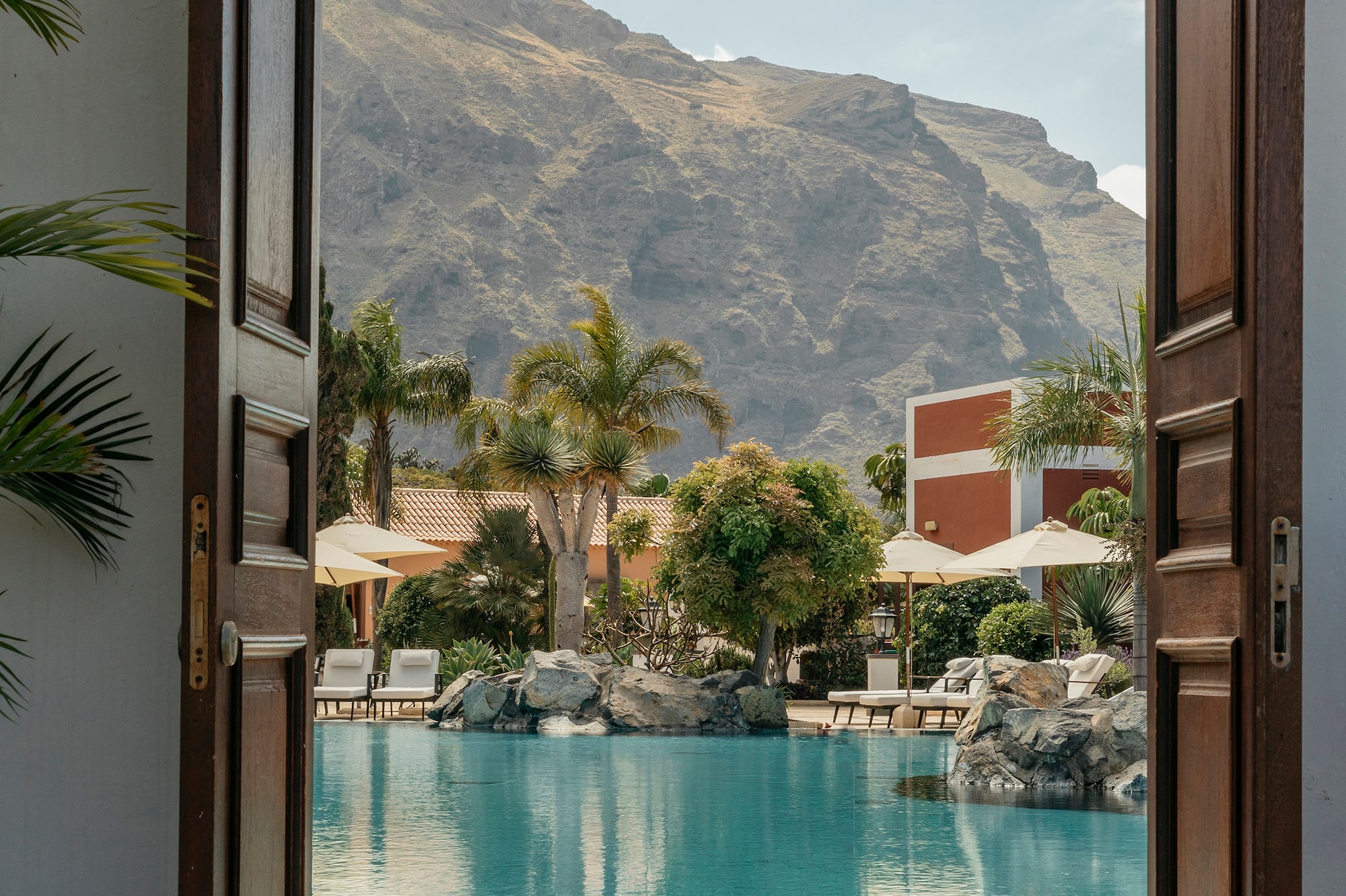Tropical resort pool with mountain backdrop framed by open doors.