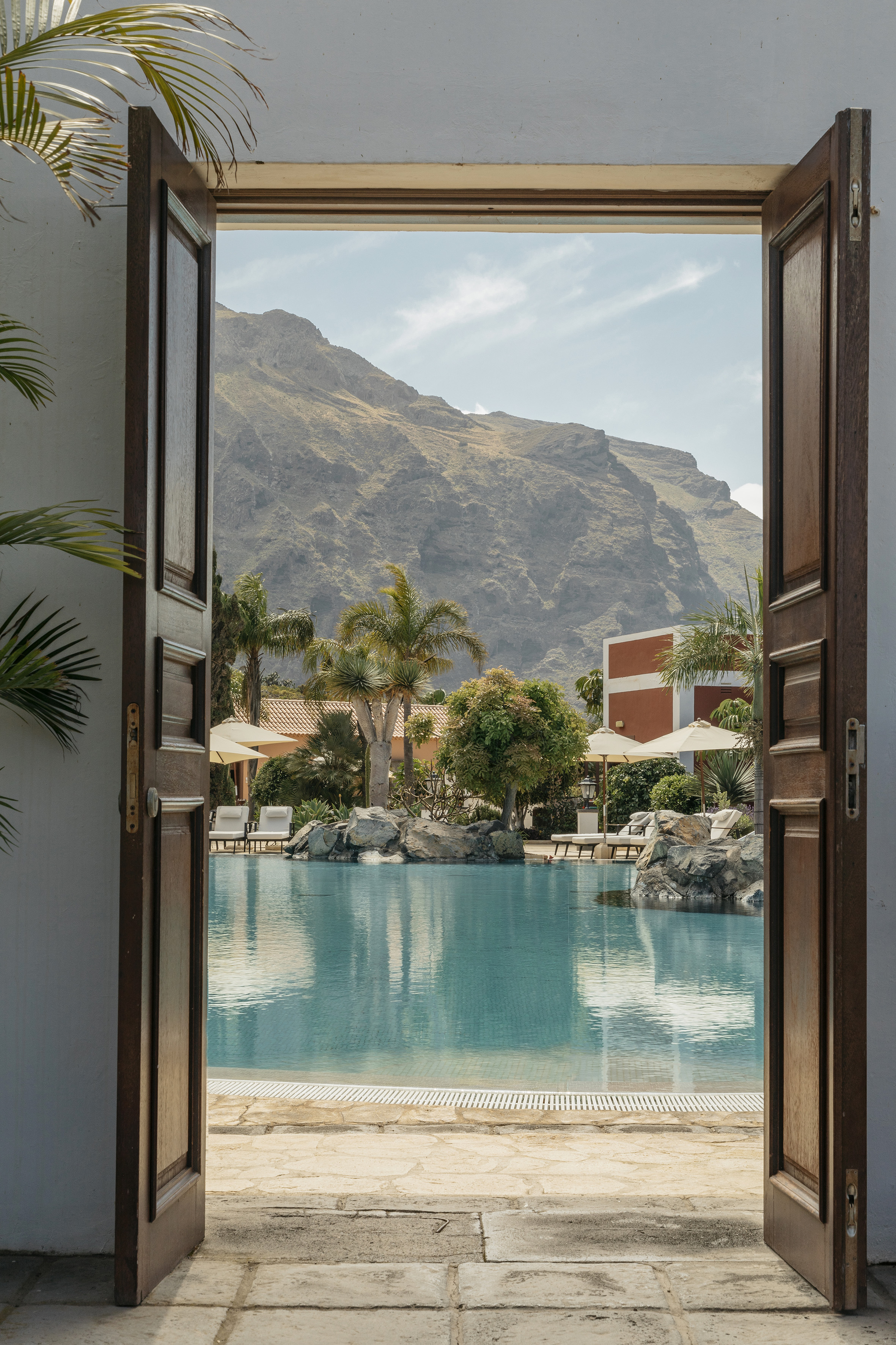 Tropical resort pool with mountain backdrop framed by open doors.
