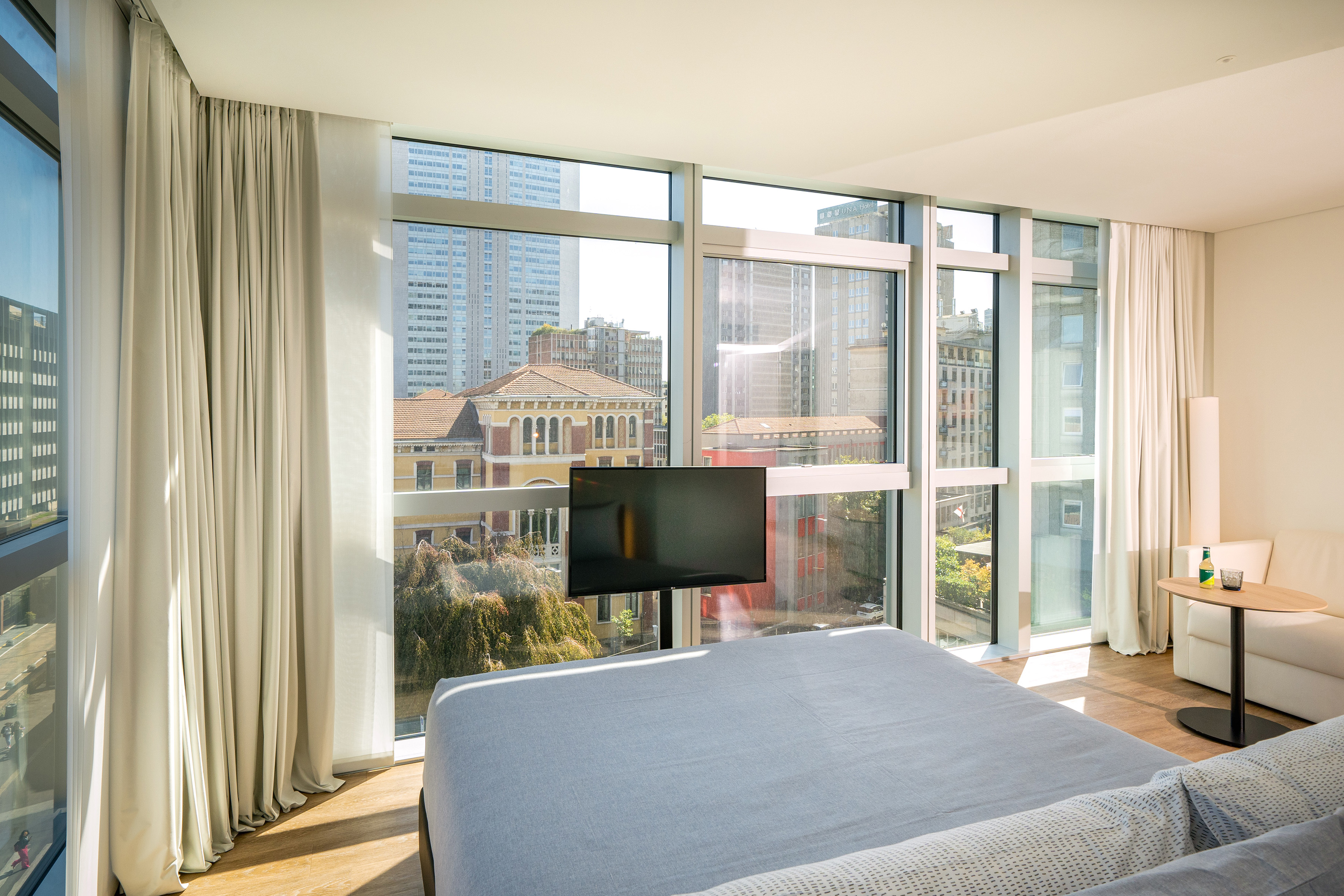 Modern hotel bedroom with panoramic city view, large windows, and TV.