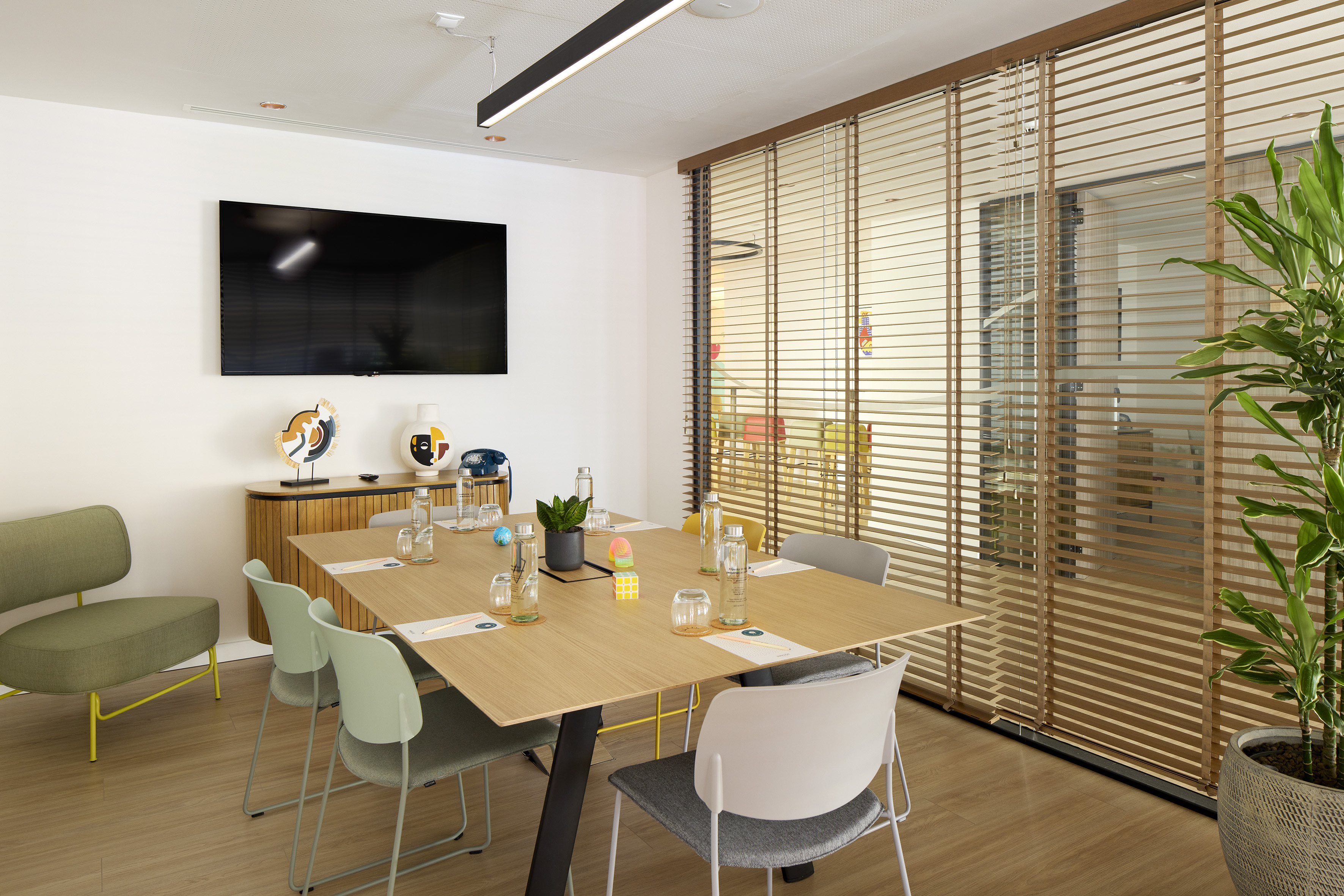 Modern meeting room with large screen, wooden table, colorful chairs, and blinds.