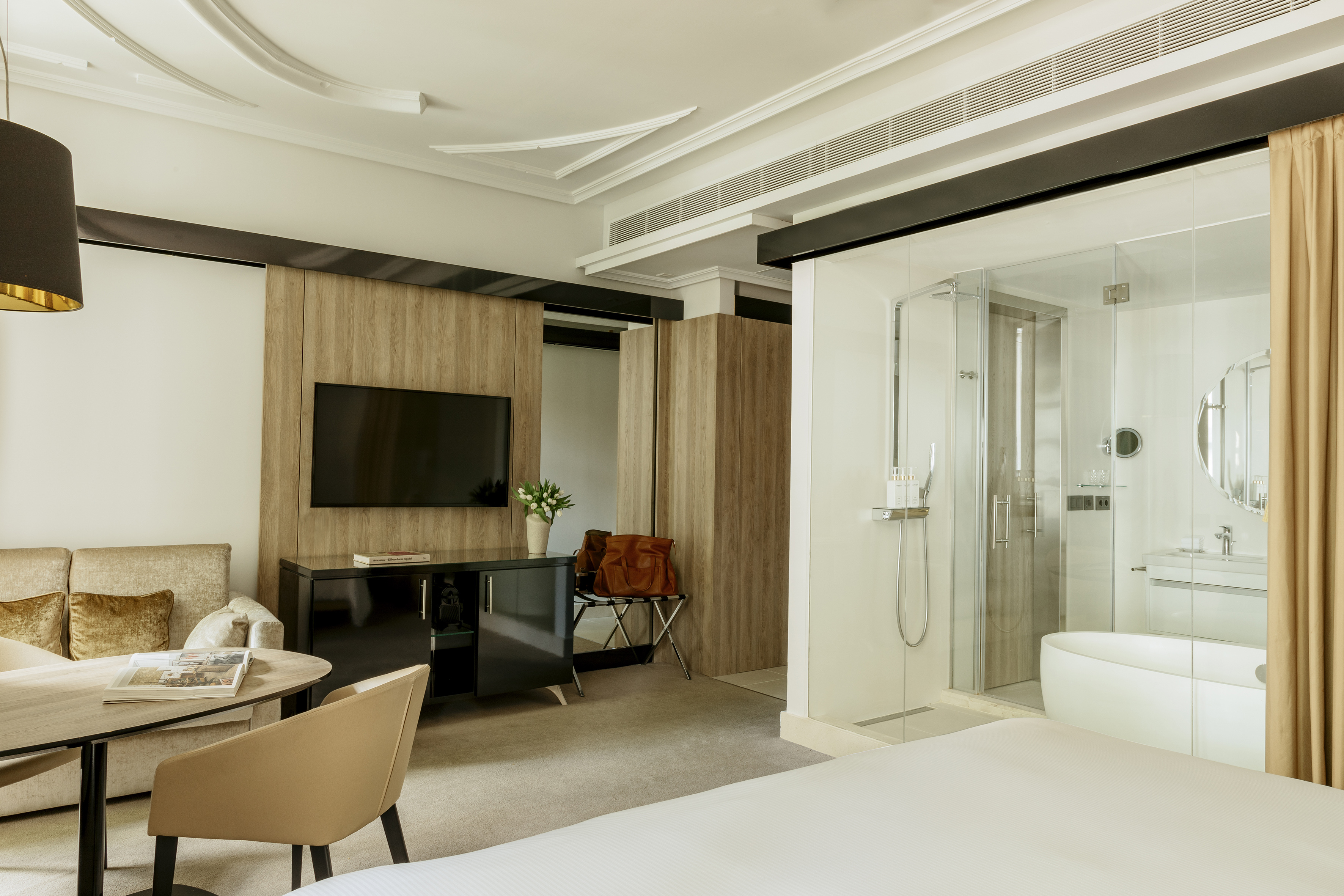 Contemporary hotel suite with bed, lounge, TV, and glass-walled bathroom.