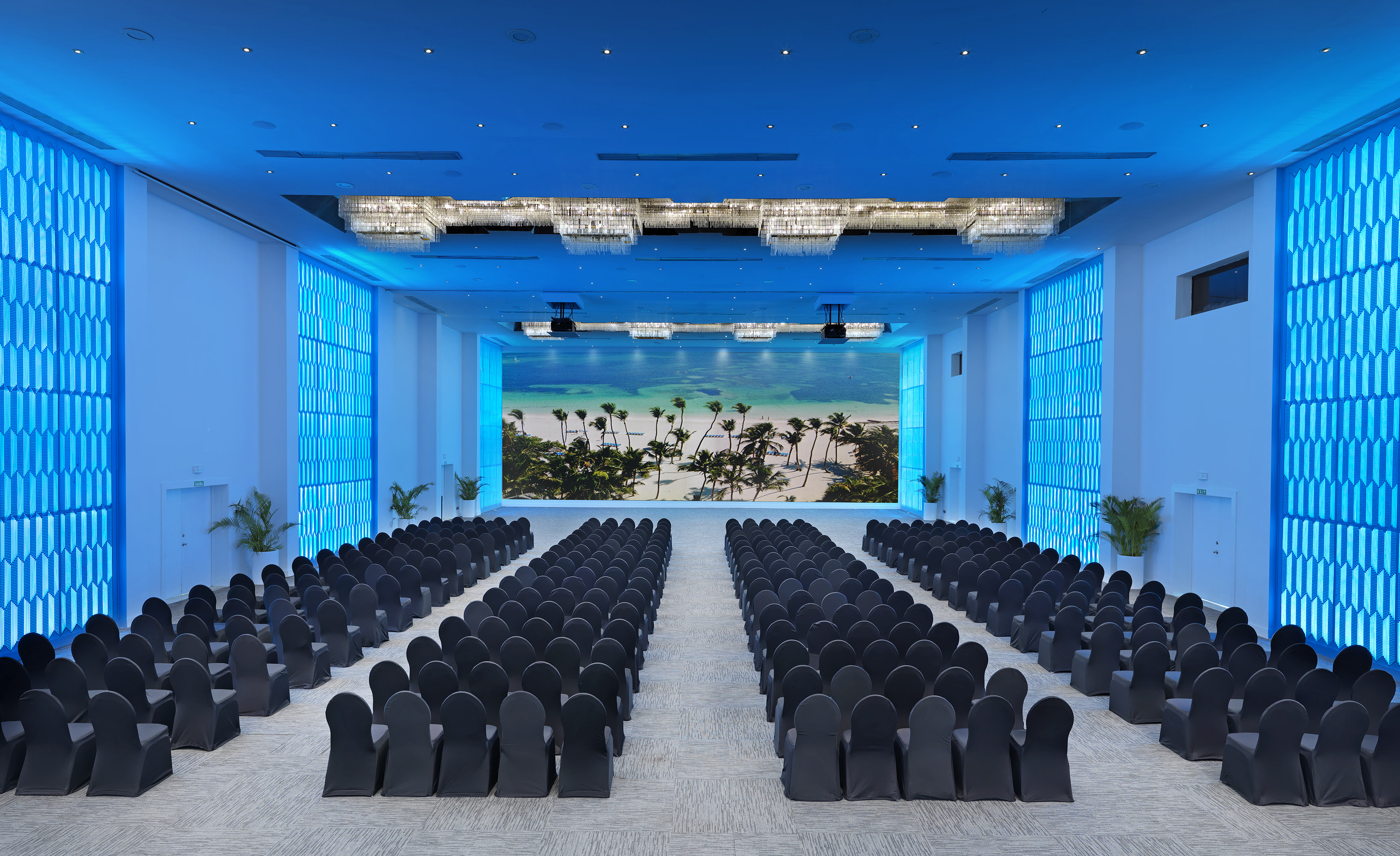 Modern event hall with theater seating, blue lighting, and large tropical beach screen.