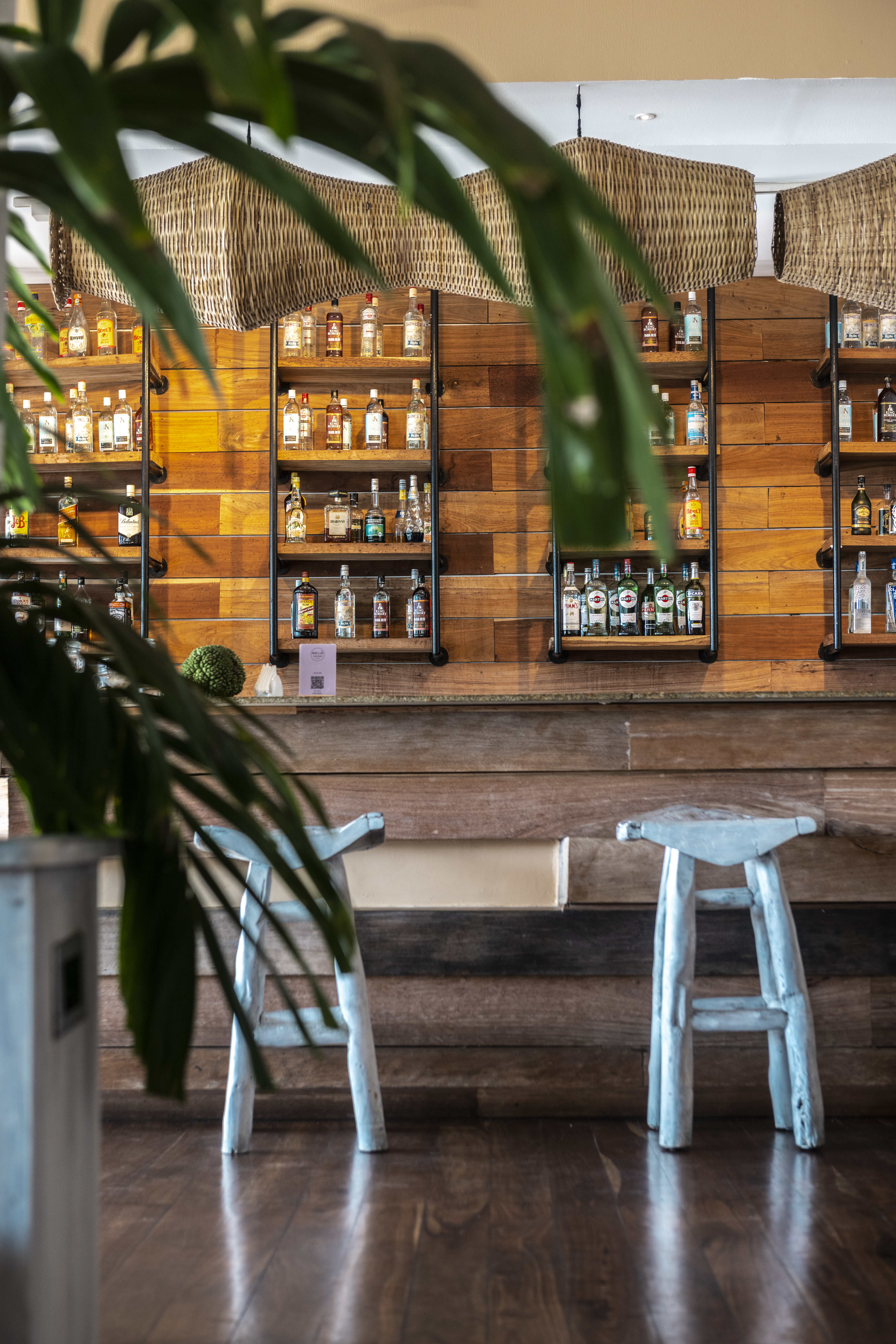 Rustic wooden bar with liquor shelves, two light stools, and wicker lighting.