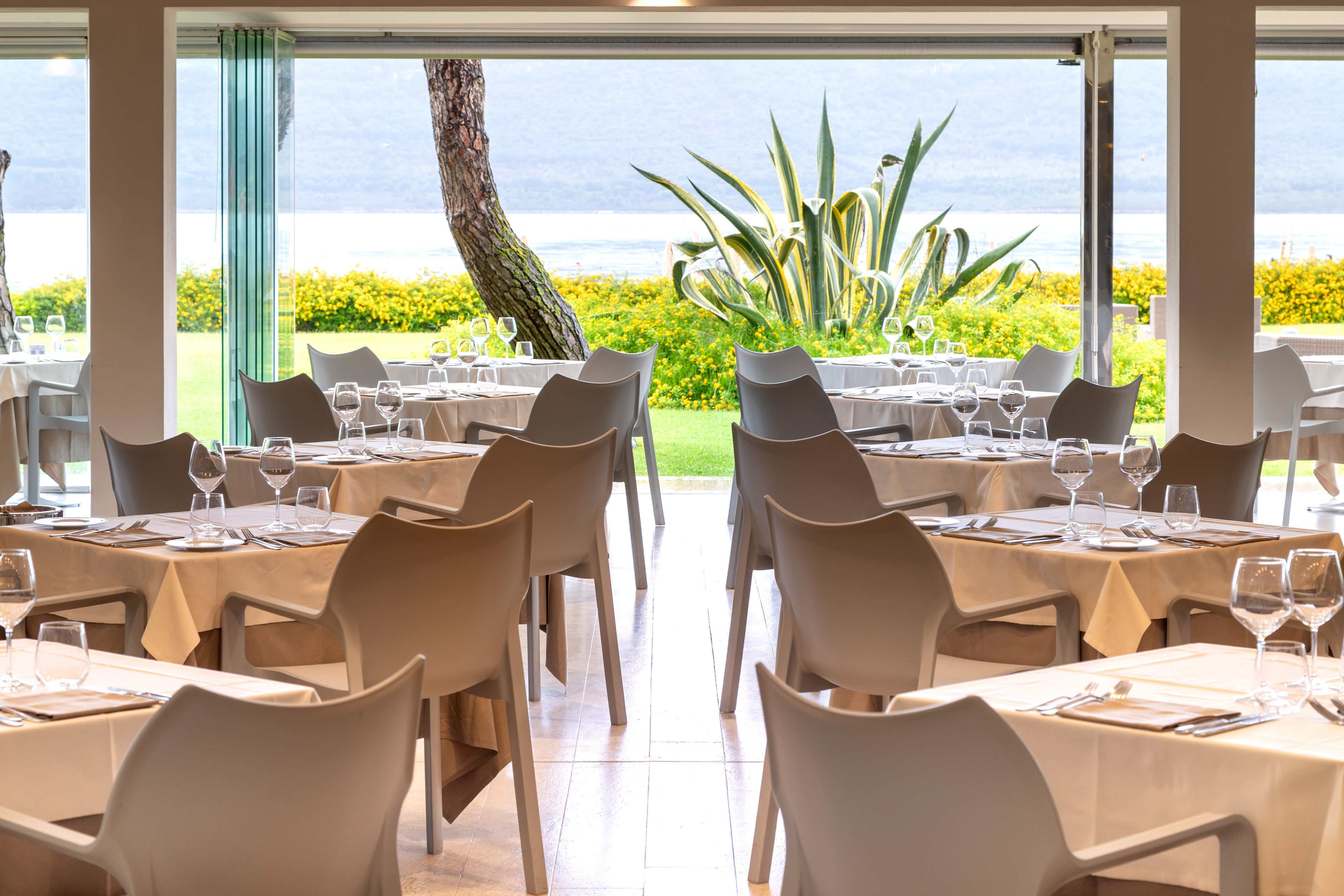 Lakeside restaurant dining room with set tables, view of water and lush plants.