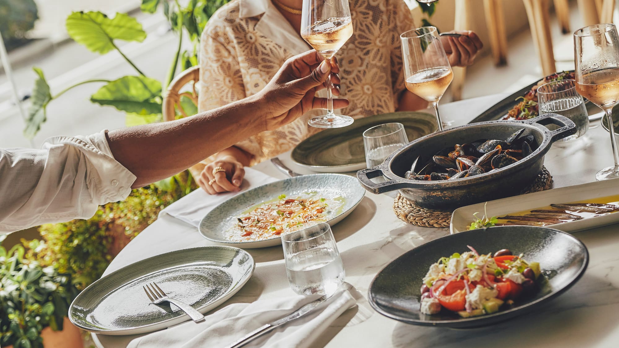 Friends dining at an ocean-view restaurant, pouring rosé wine with mussels and salads.