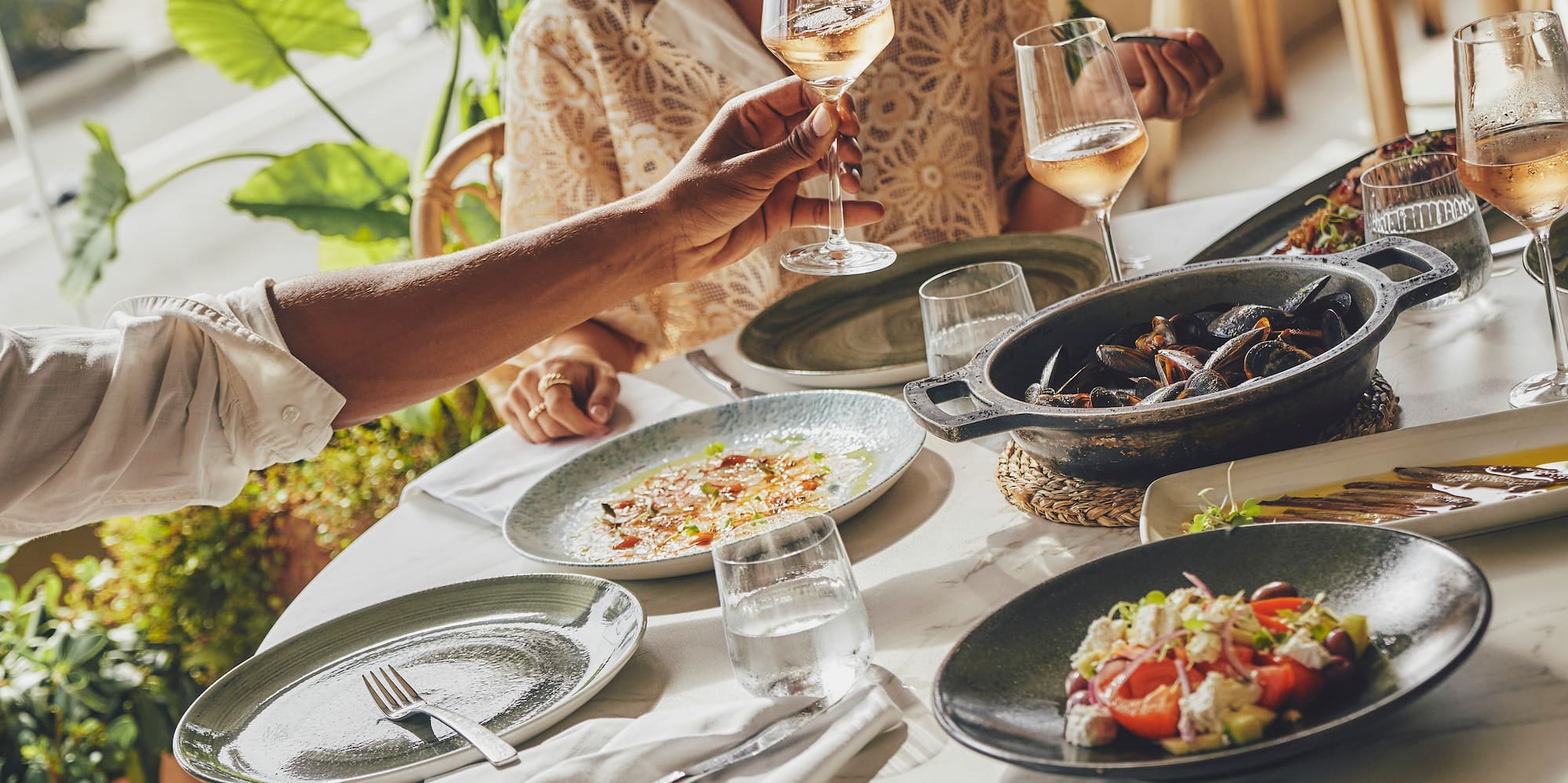 Friends dining at an ocean-view restaurant, pouring rosé wine with mussels and salads.