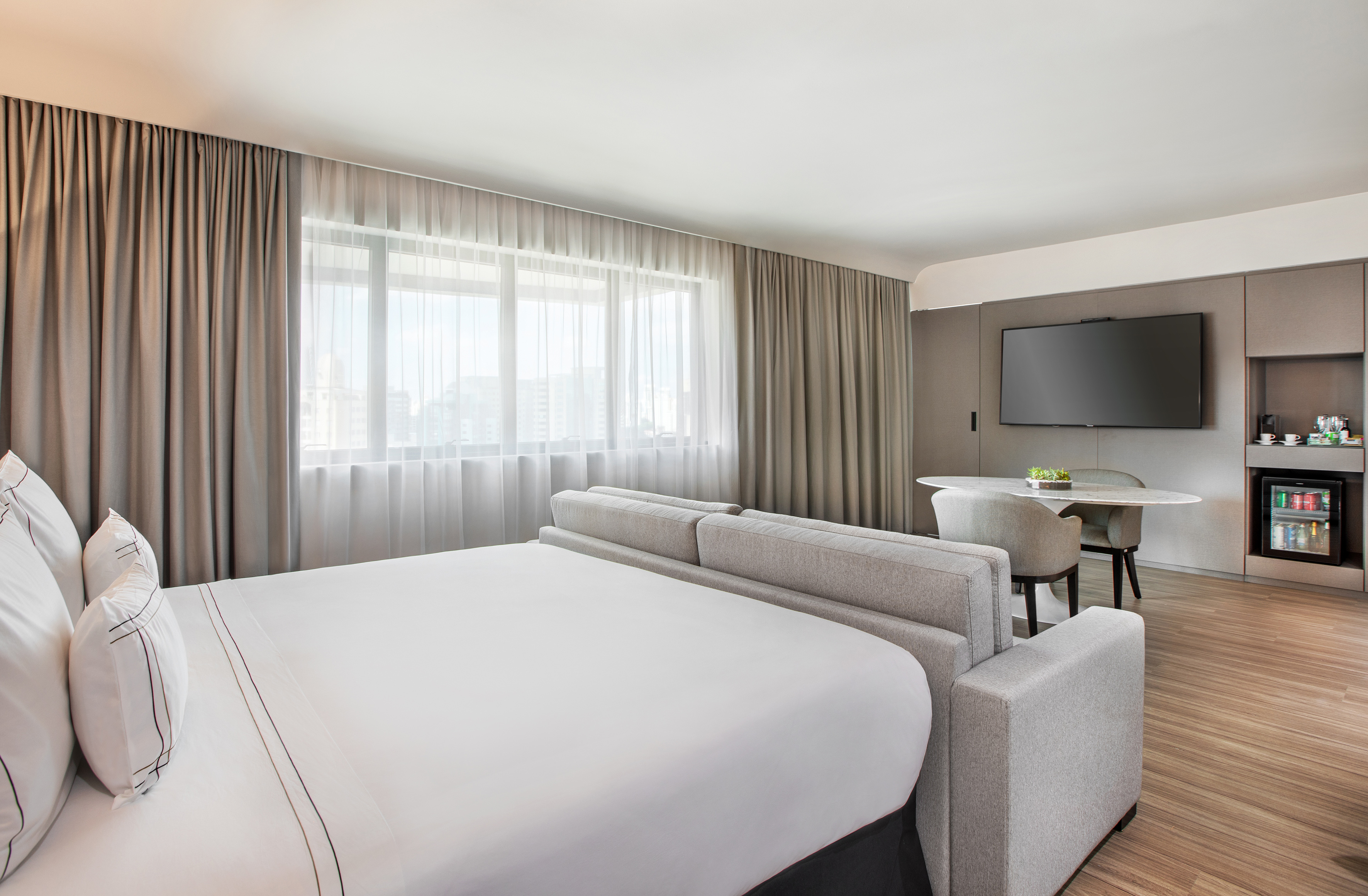 Modern hotel room with king bed, sofa, dining area, wall TV, and minibar.