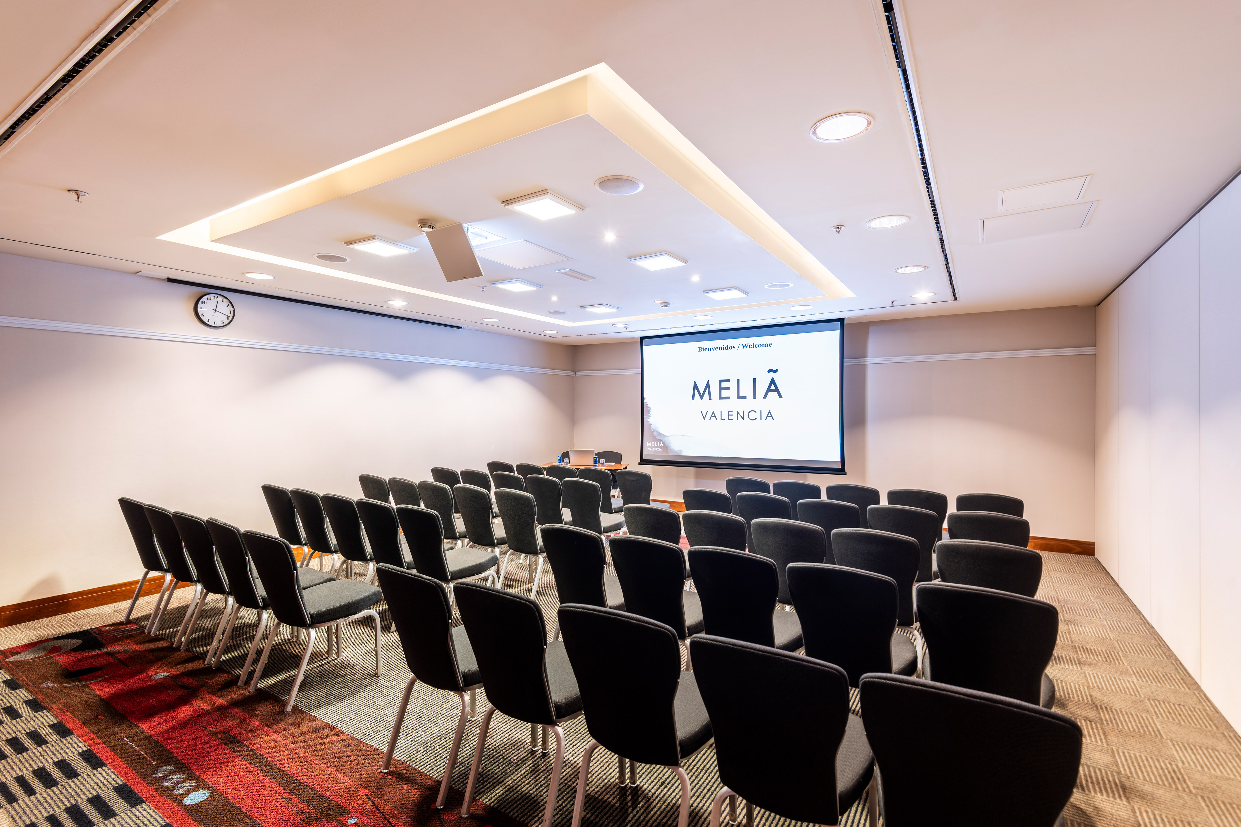 Empty hotel meeting room with black chairs facing a Meliã Valencia screen.