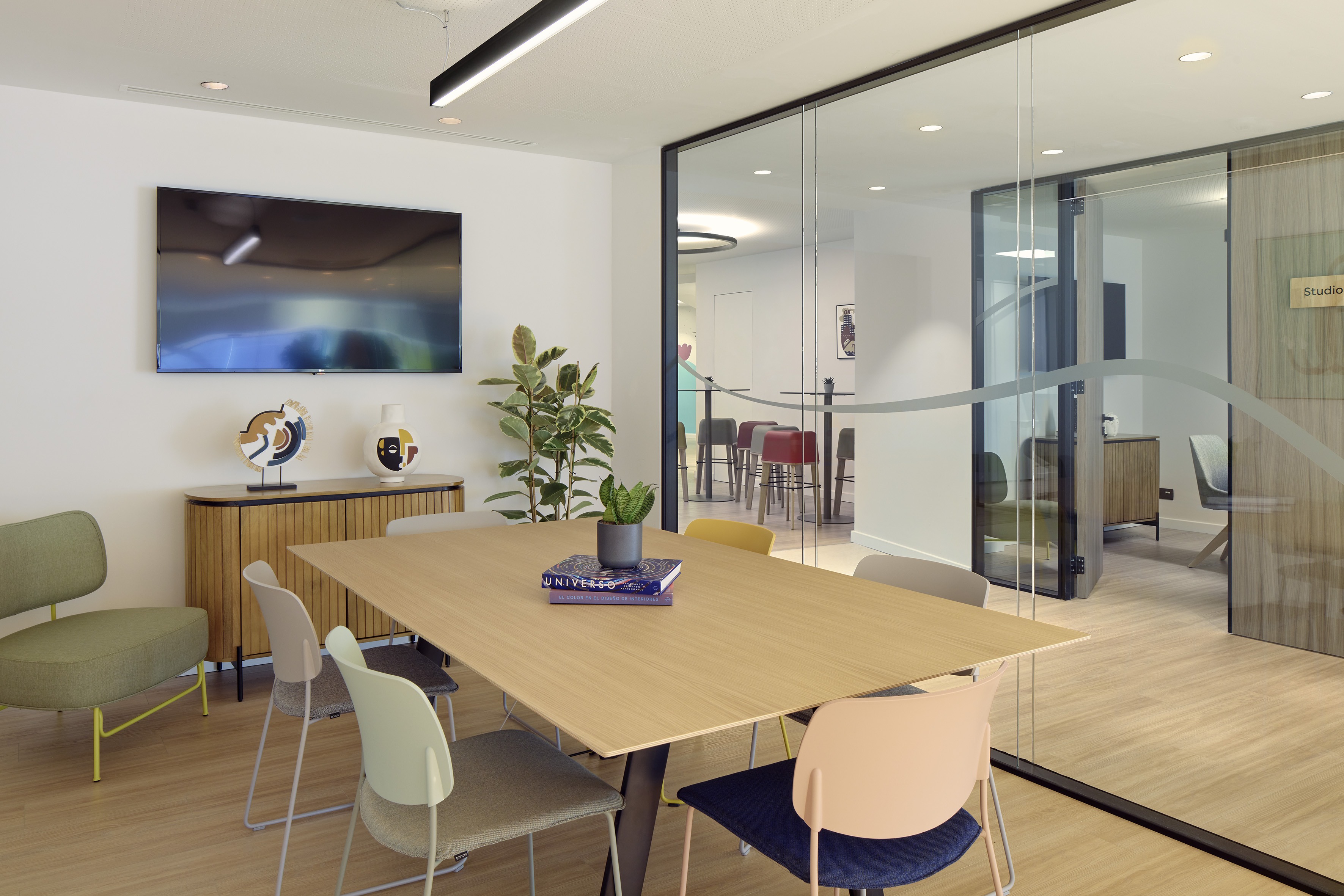 Modern office meeting room with wooden table, colorful chairs, TV, and glass wall.