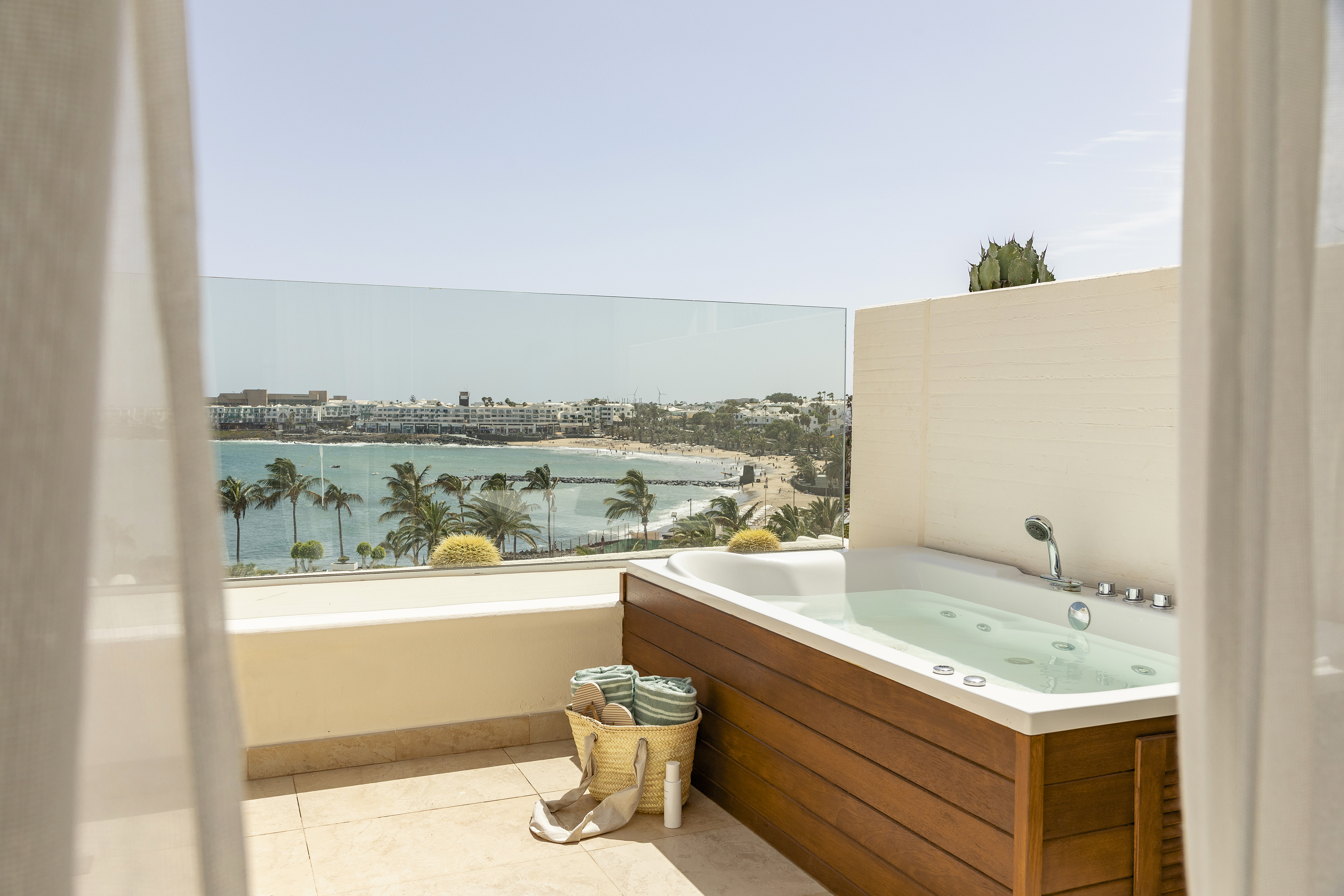 Balcony jacuzzi with sea view, sandy beach, palm trees, and resort.