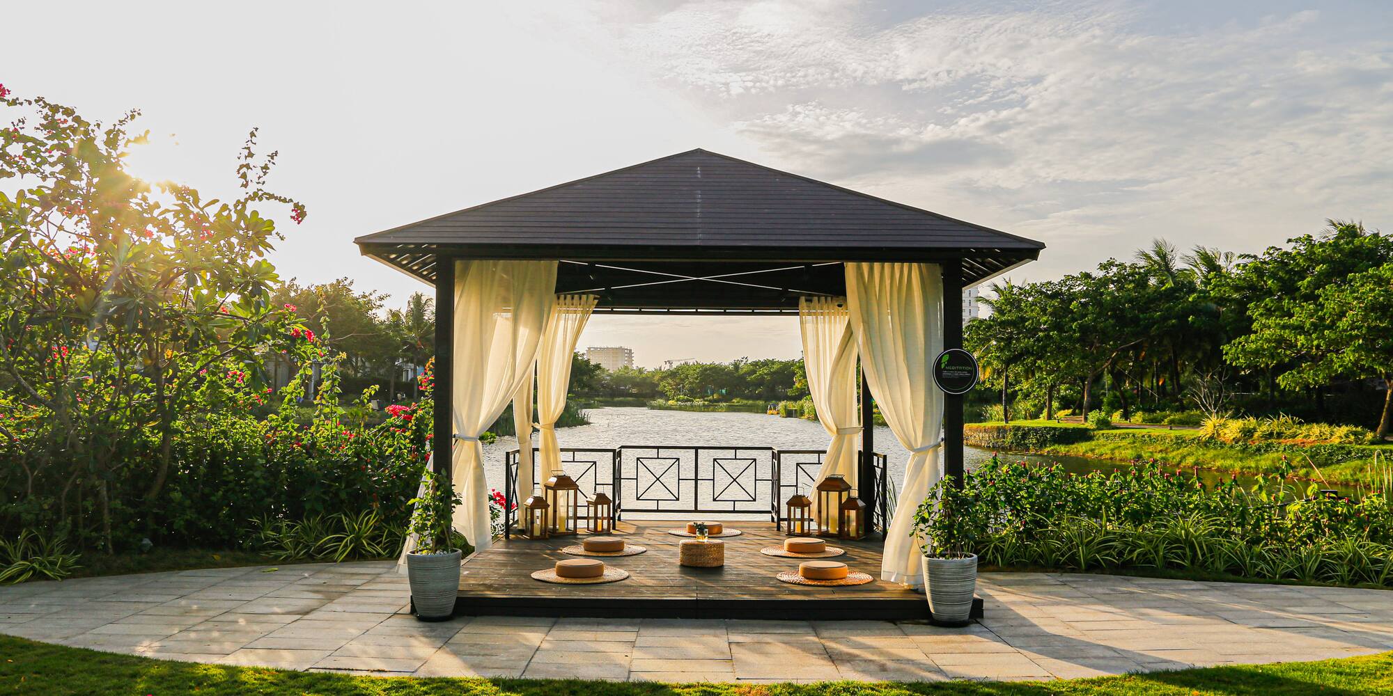 Waterfront gazebo with lanterns, cushions, and lush gardens at sunset.
