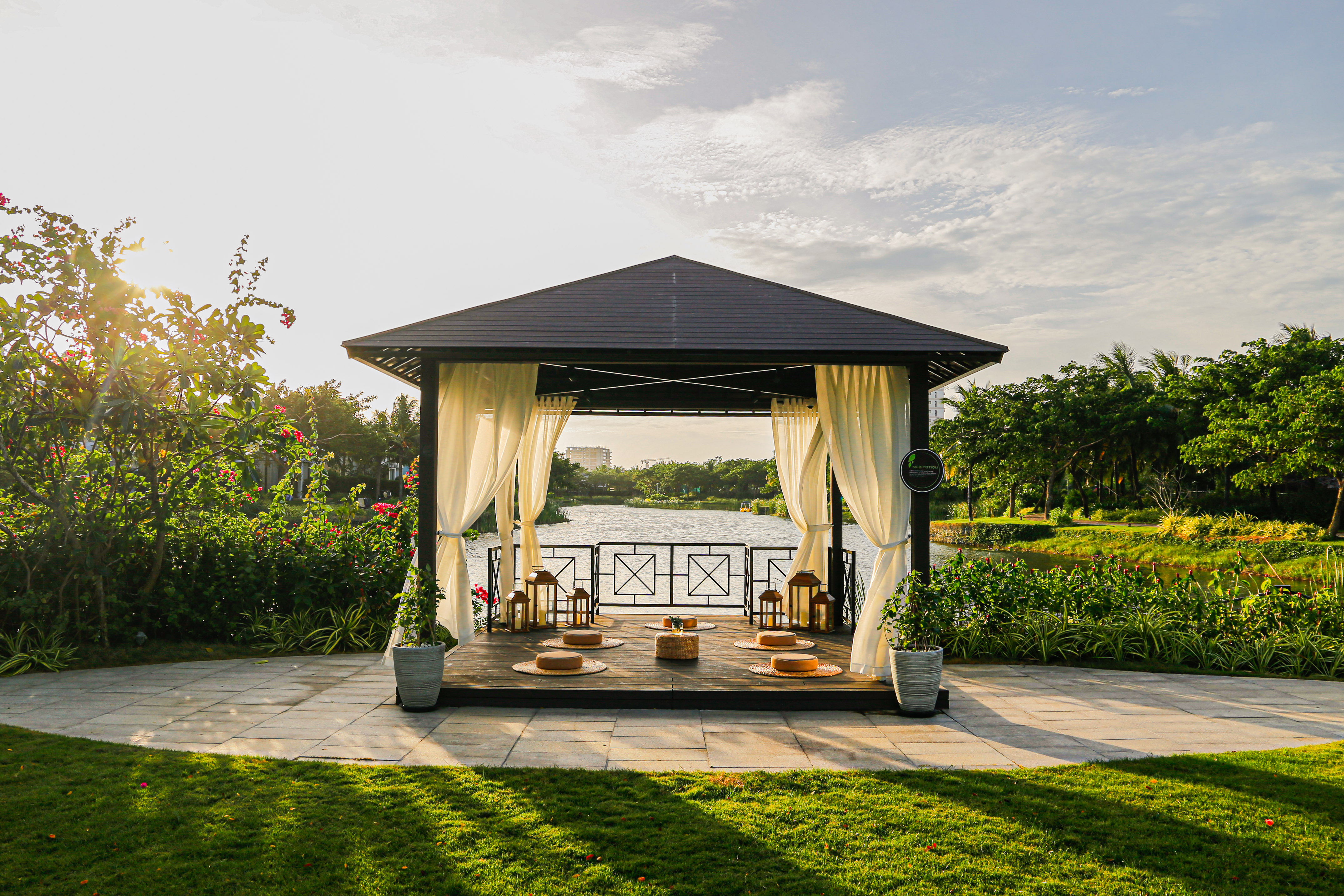 Waterfront gazebo with lanterns, cushions, and lush gardens at sunset.