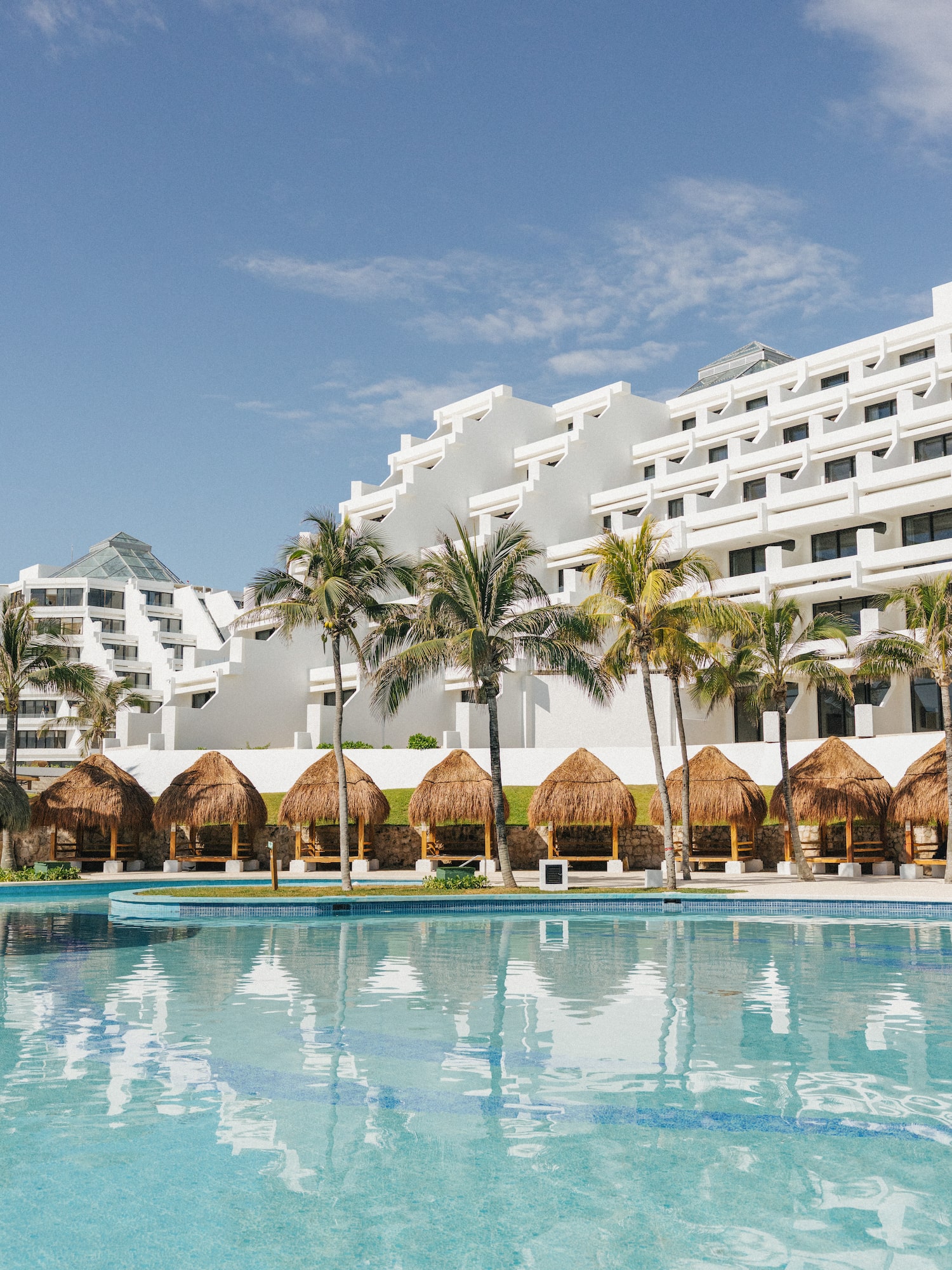 Modern resort hotel, blue pool, palm trees, and thatched cabanas under sunny sky.