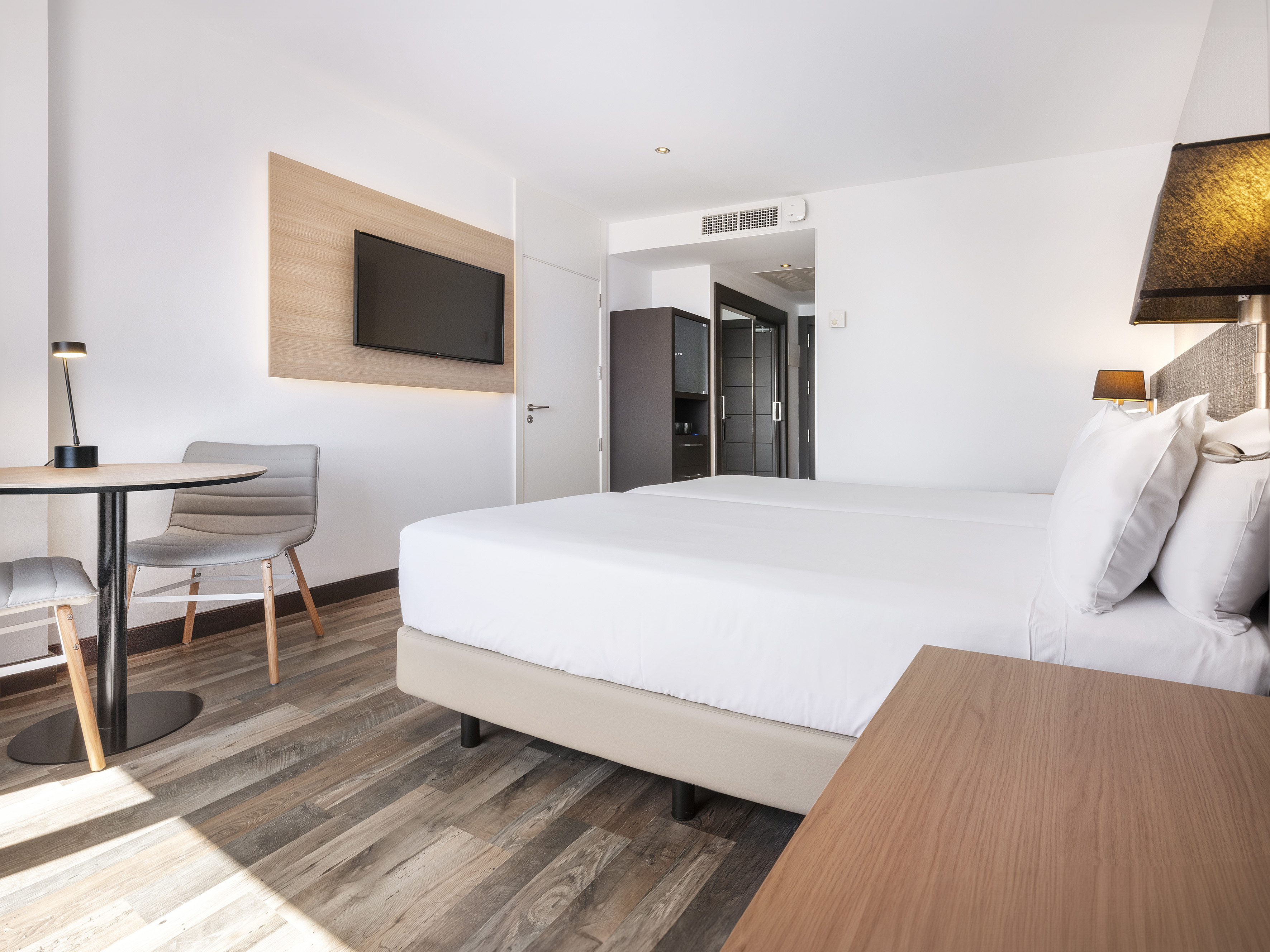 Modern hotel room: twin beds, wall TV, small table, and chairs.