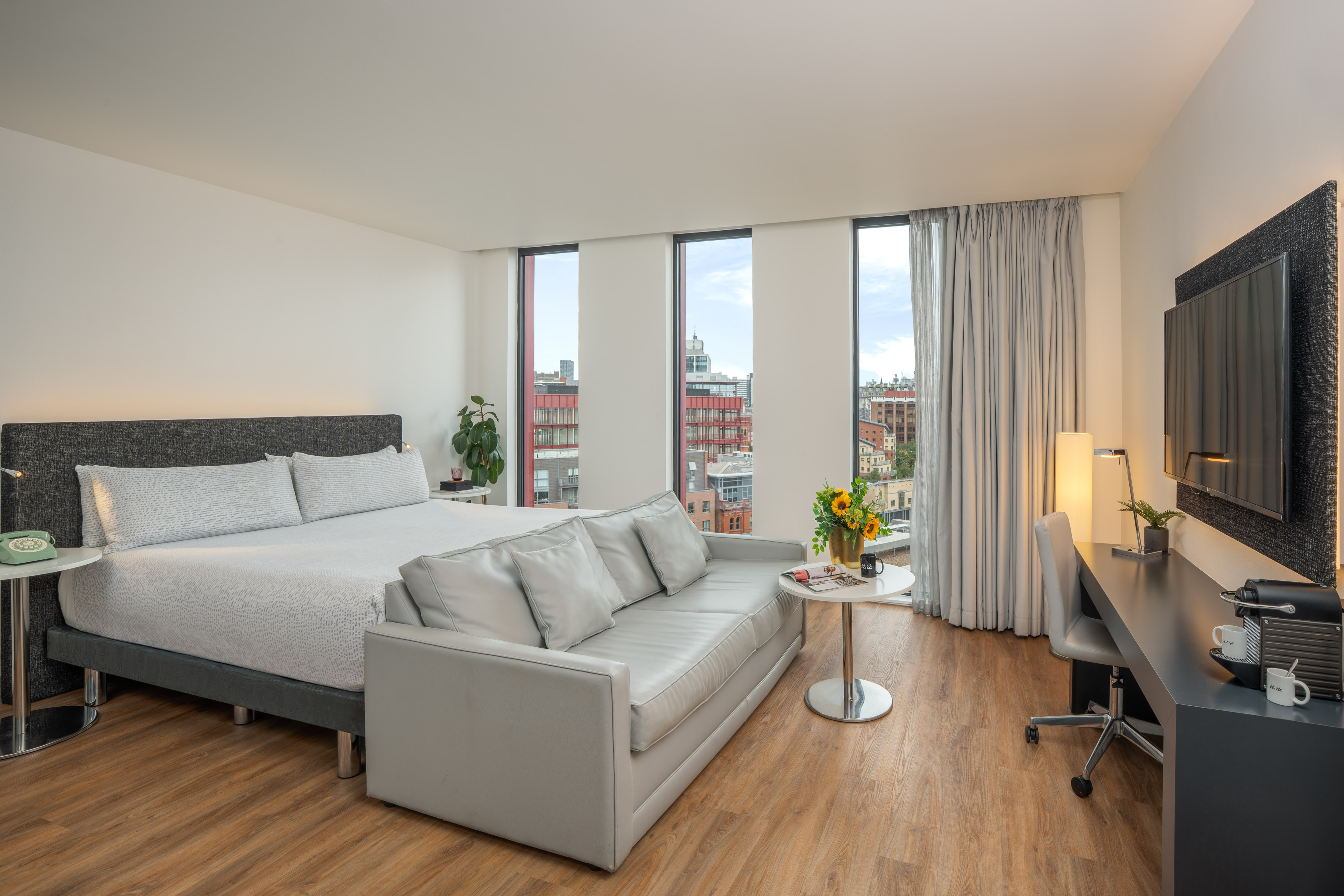 Hotel room with king bed, grey sofa, desk, large windows, and city view.