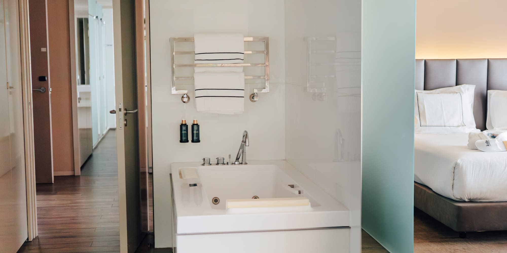Modern hotel suite bathroom with white bathtub, towels, and adjacent bedroom.