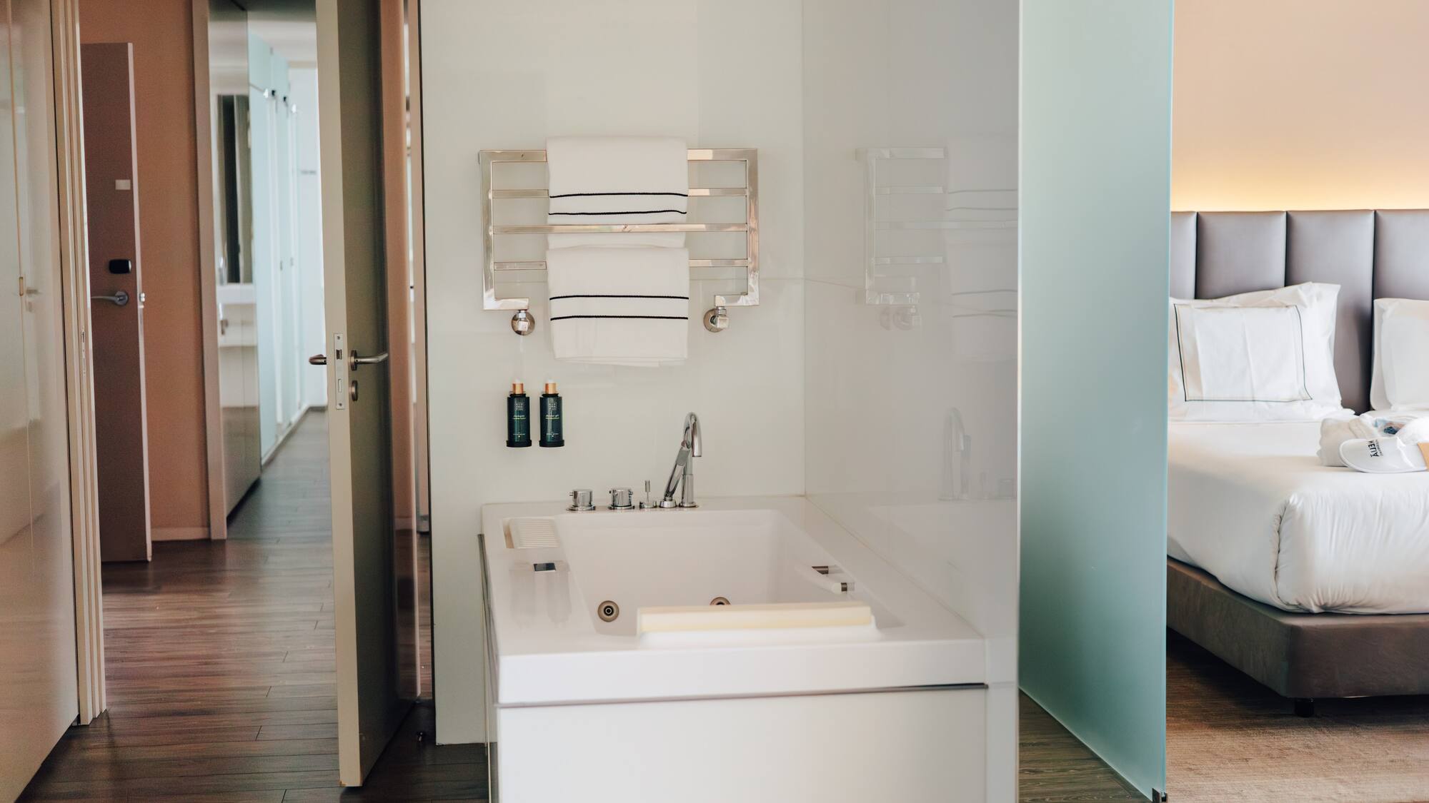 Modern hotel suite bathroom with white bathtub, towels, and adjacent bedroom.