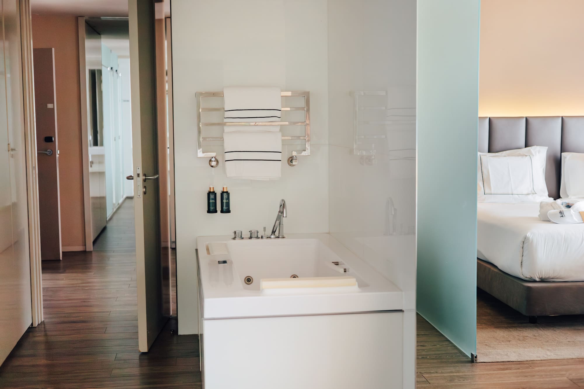 Modern hotel suite bathroom with white bathtub, towels, and adjacent bedroom.