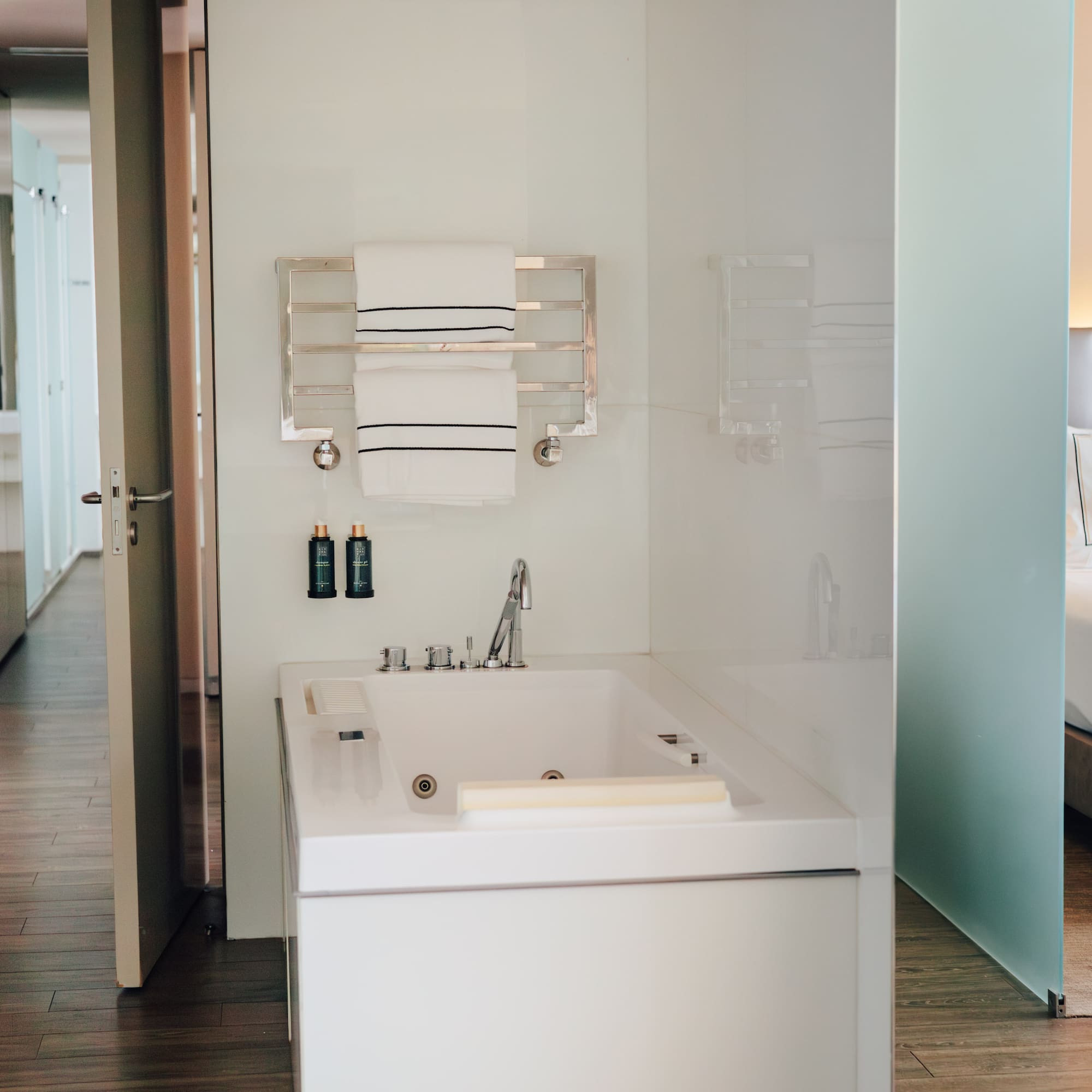 Modern hotel suite bathroom with white bathtub, towels, and adjacent bedroom.