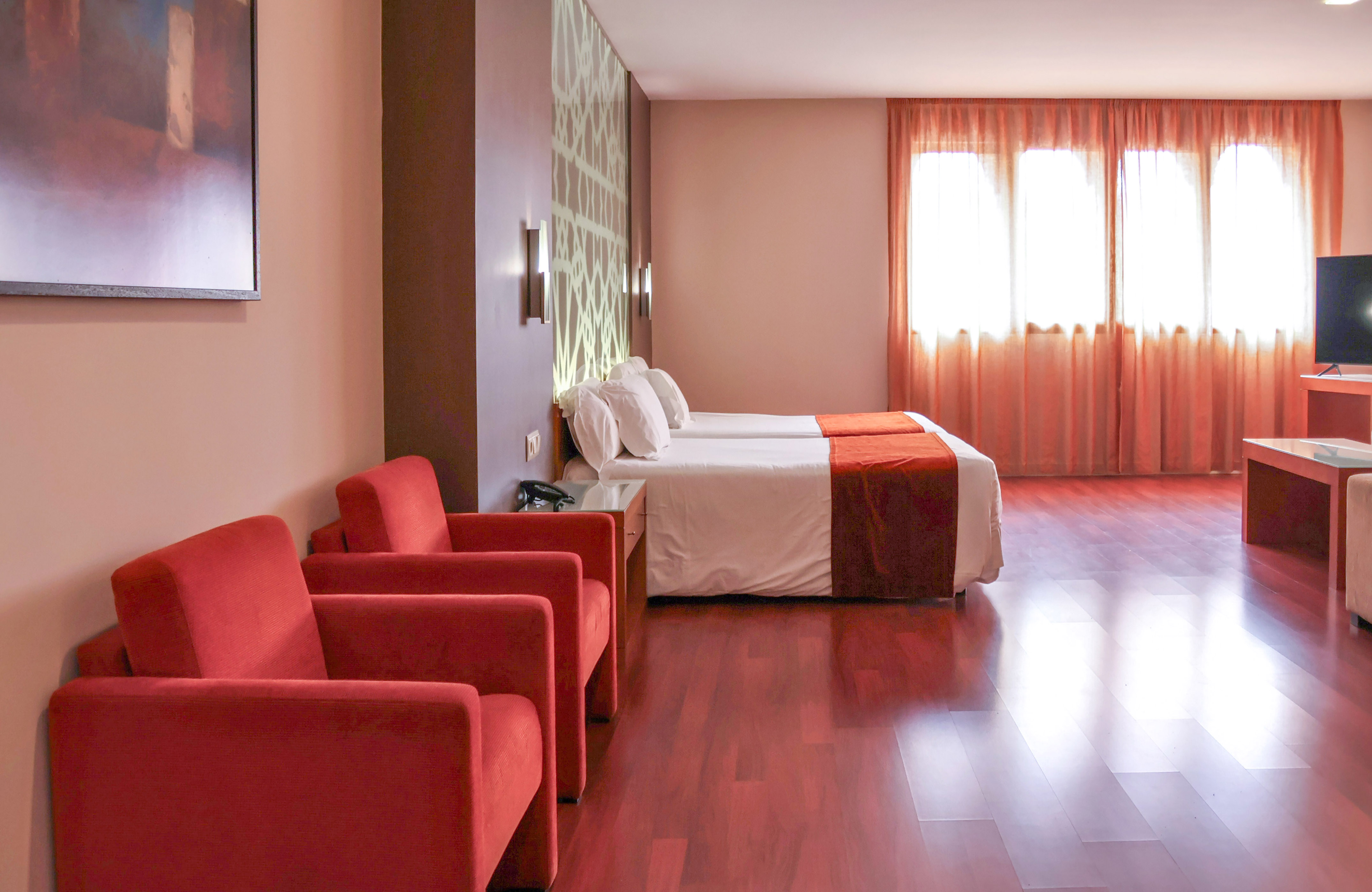 Modern hotel room with twin beds, red armchairs, and wood floor.
