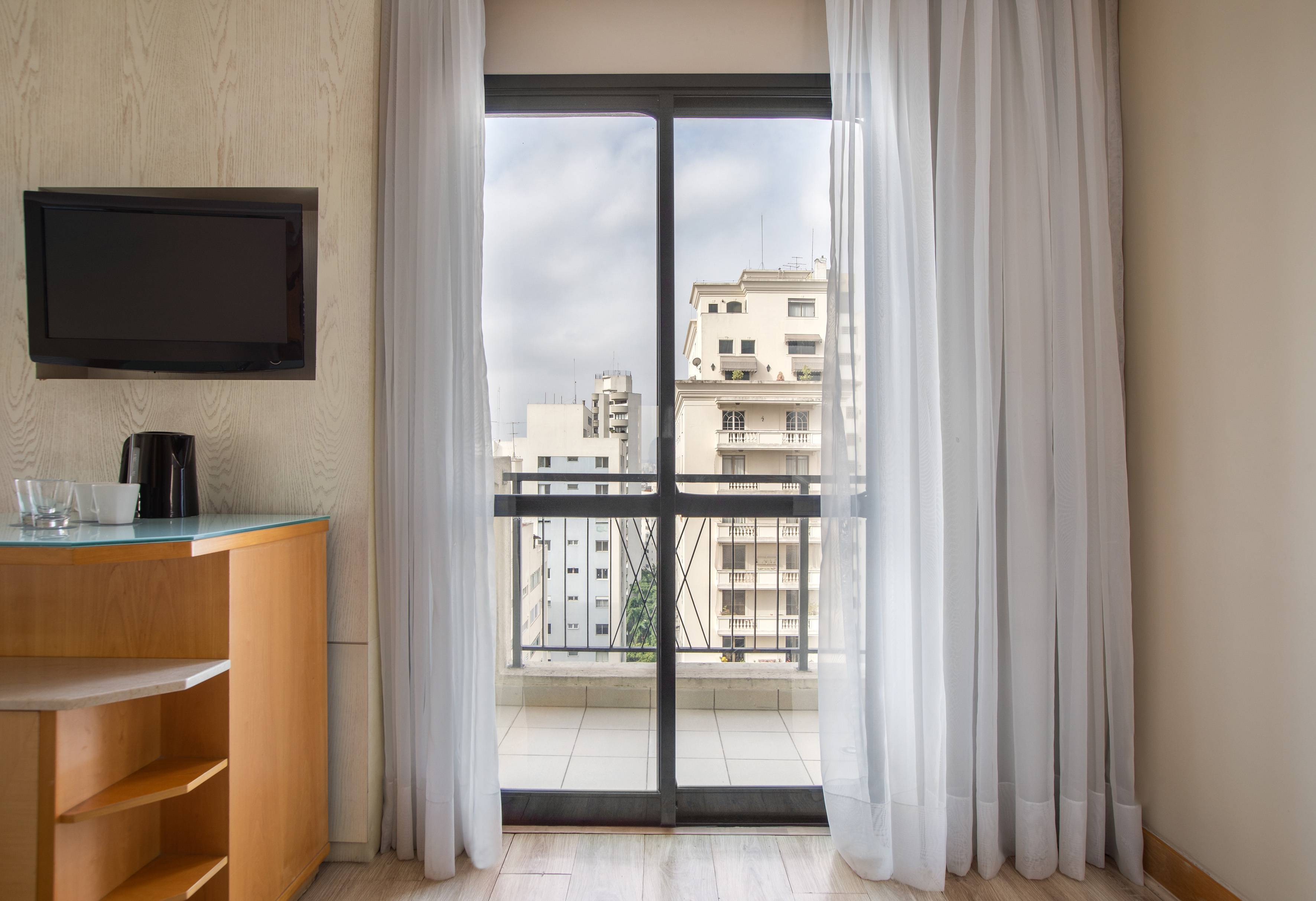 Modern hotel room with city view balcony, white curtains, TV, and cabinet.