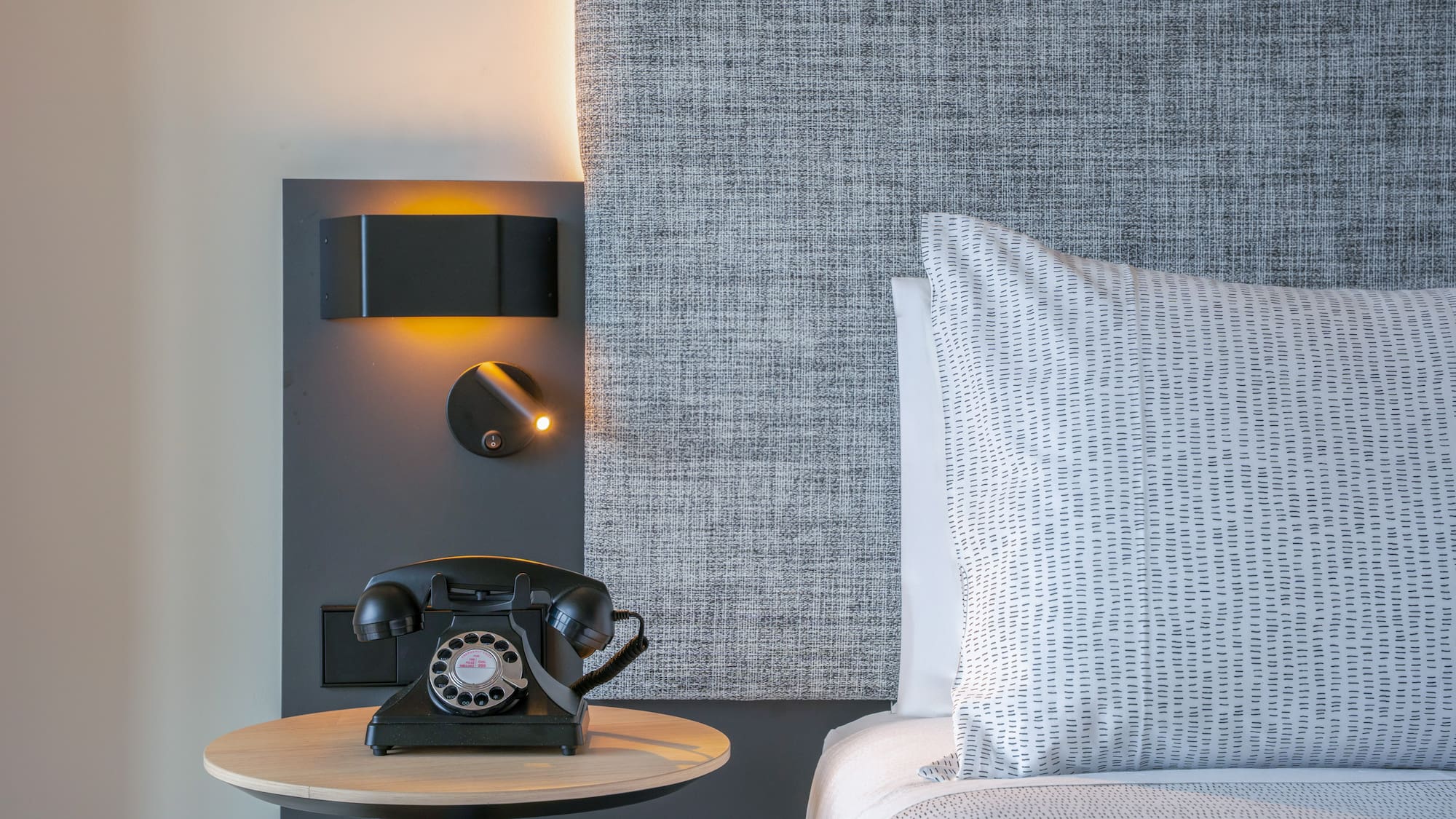 Dormitorio de hotel con teléfono retro, lámparas y cabecero gris.