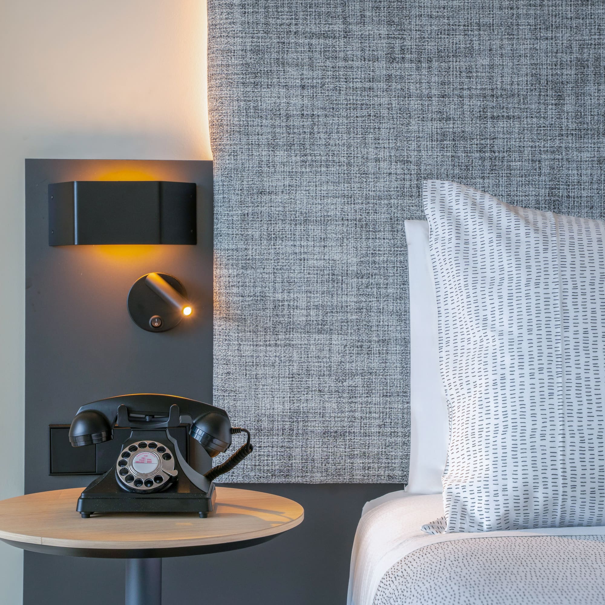 Dormitorio de hotel con teléfono retro, lámparas y cabecero gris.