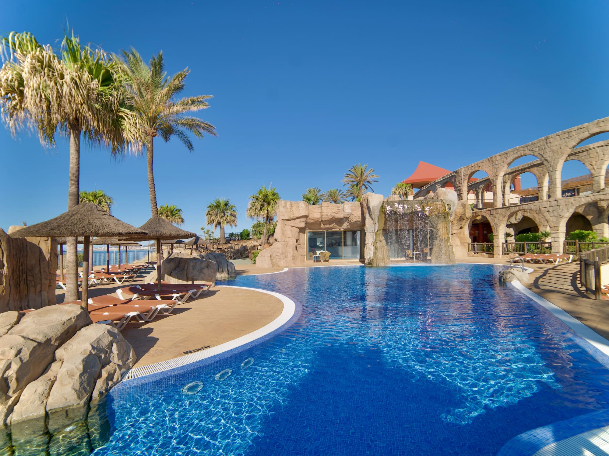 Piscina azul de hotel con palmeras, cascada y arcos de piedra cerca del mar.