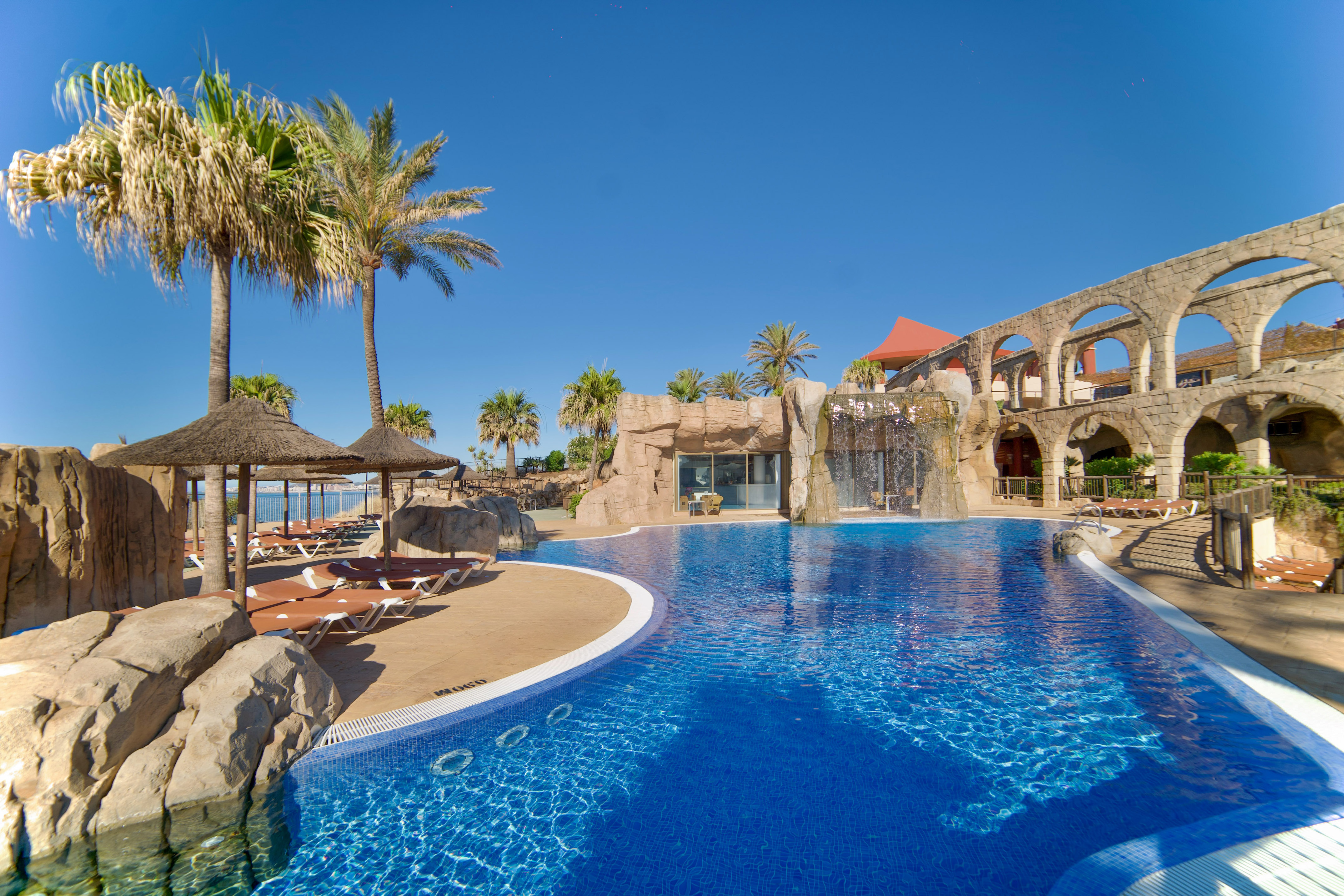 Piscina azul de hotel con palmeras, cascada y arcos de piedra cerca del mar.