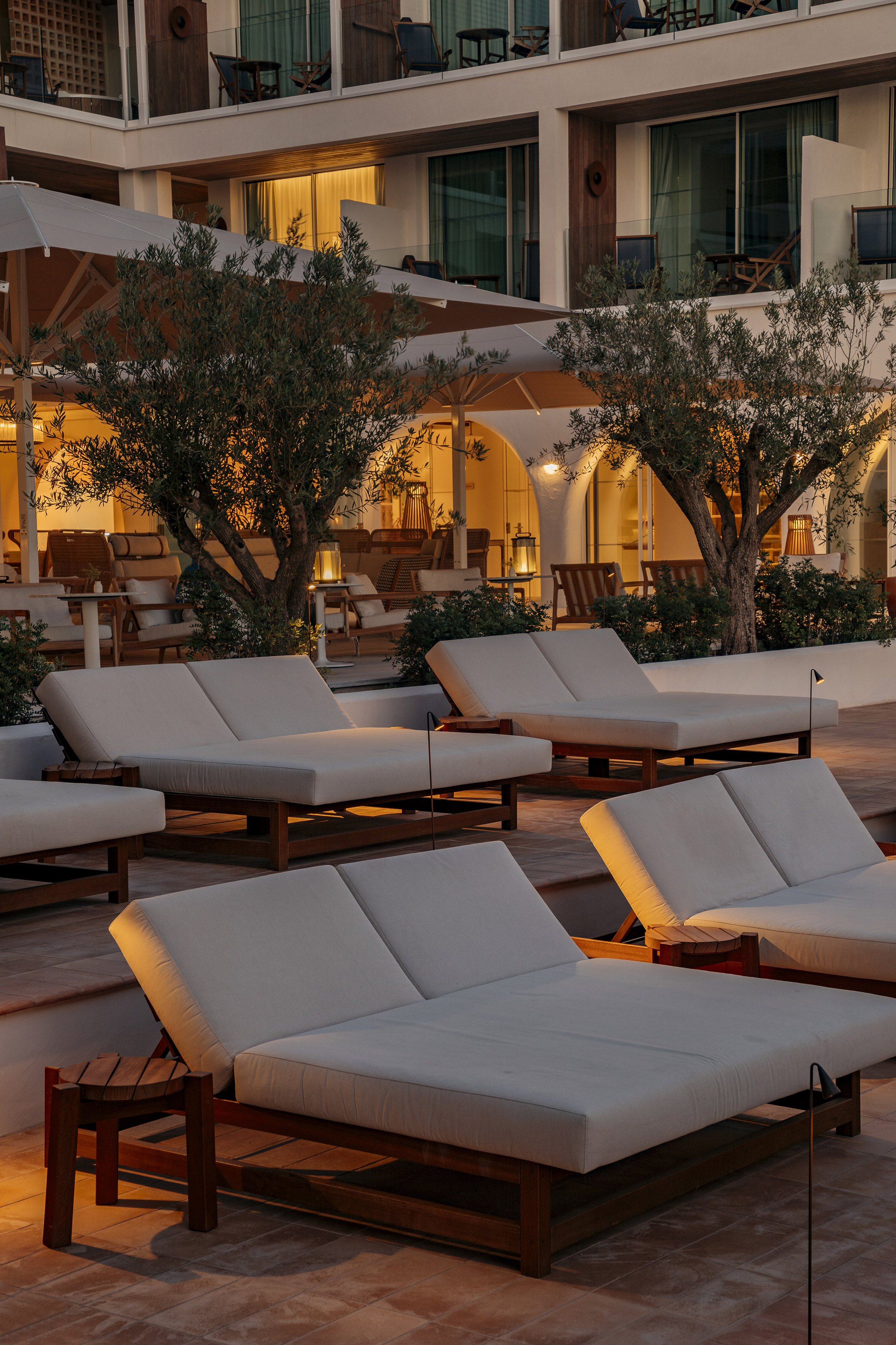 Warmly lit resort patio at dusk with white sun loungers and olive trees.
