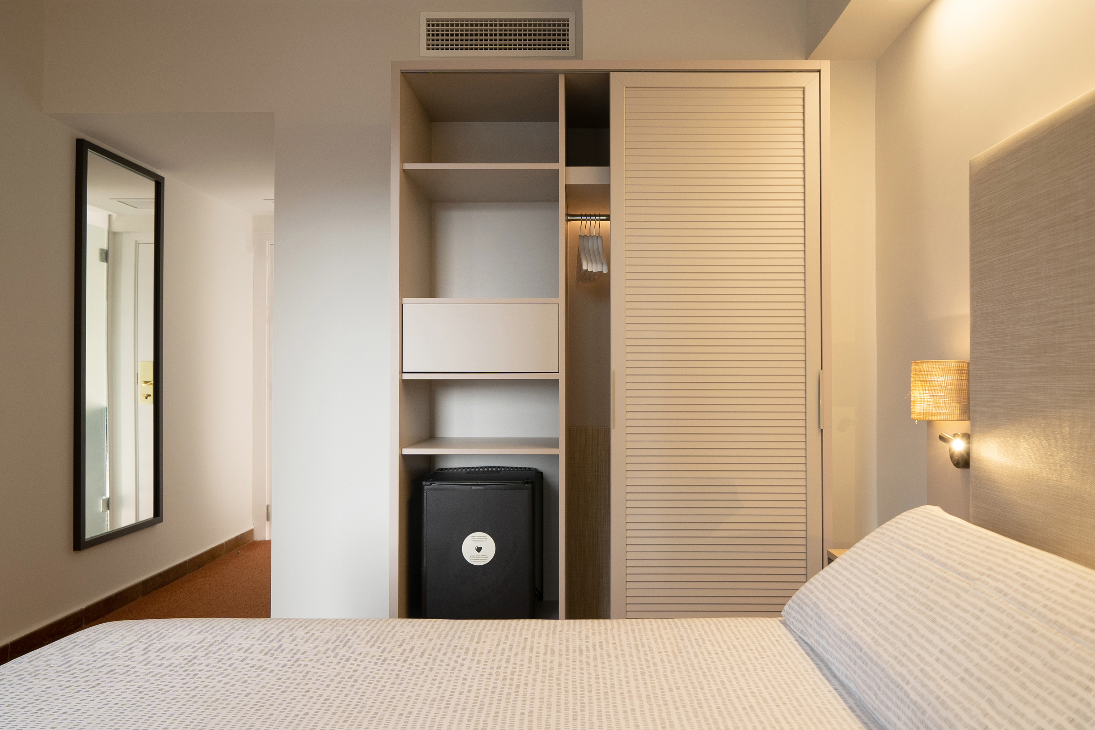 Hotel bedroom with light wood closet, minibar, and full-length mirror.