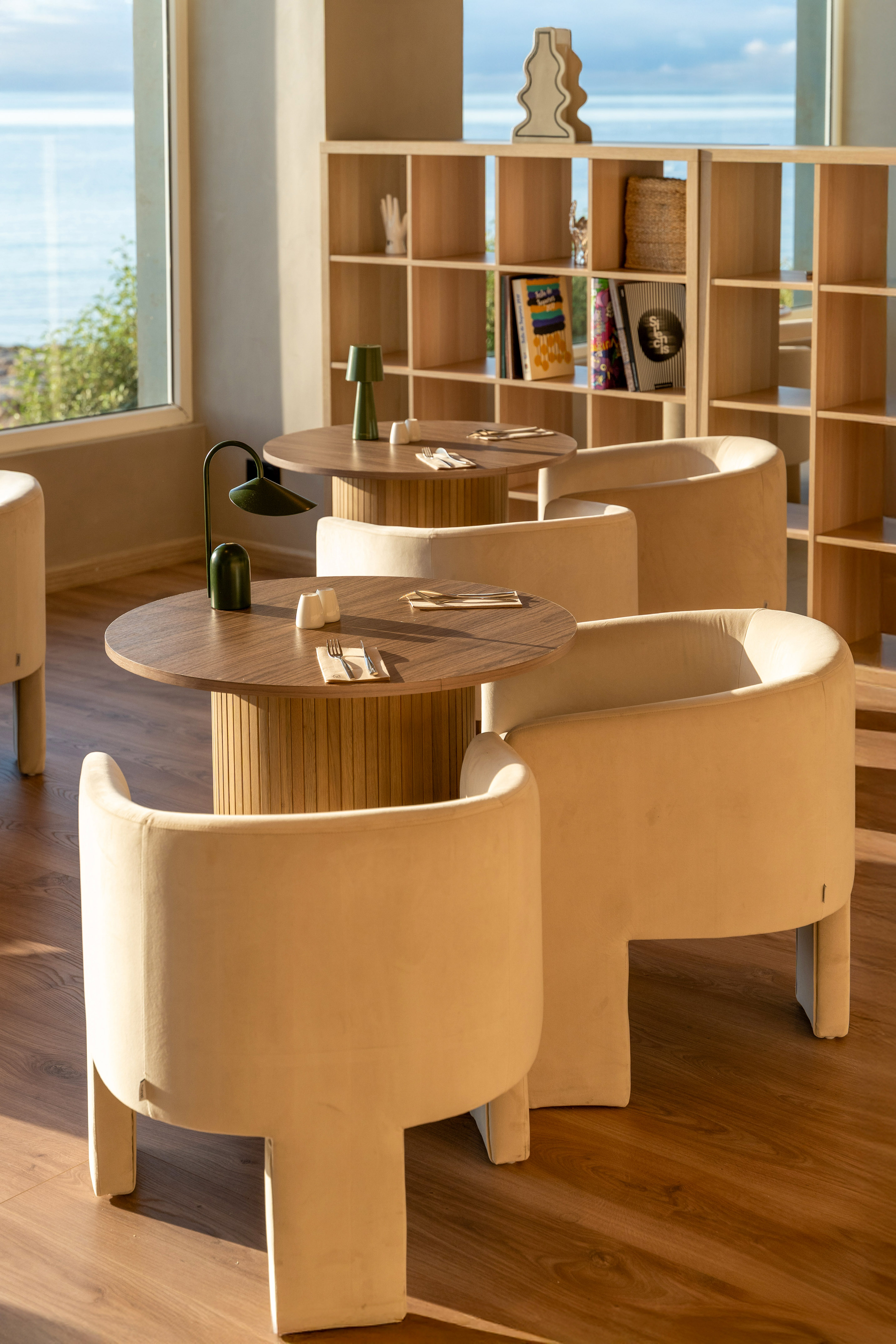 Modern coastal cafe with round wooden tables, cream chairs, ocean view, and bookshelf.