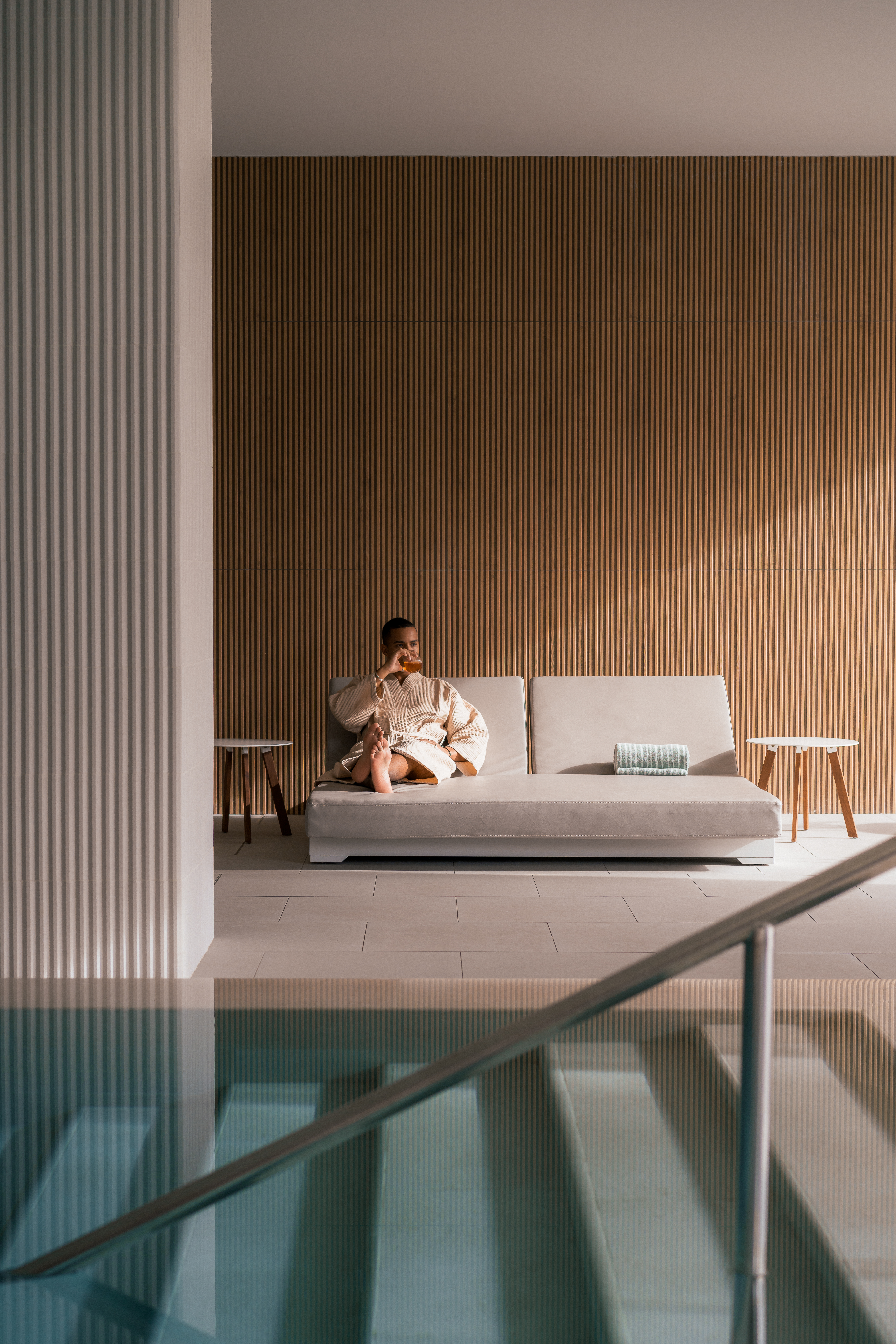 Hombre en albornoz relajándose en sofá, piscina interior spa moderno.