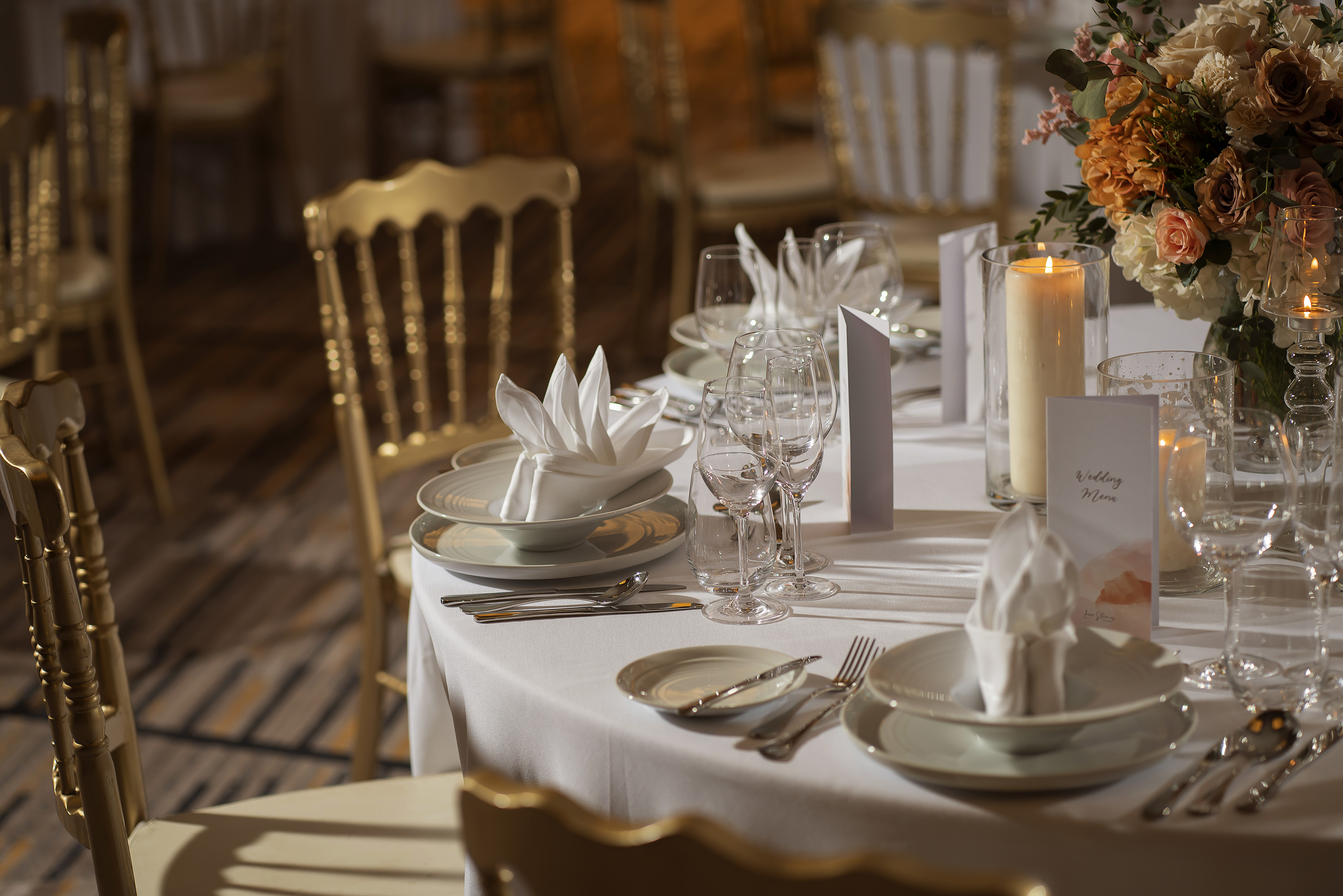 Elegant wedding table with floral centerpiece, candles, menu, and gold chairs.