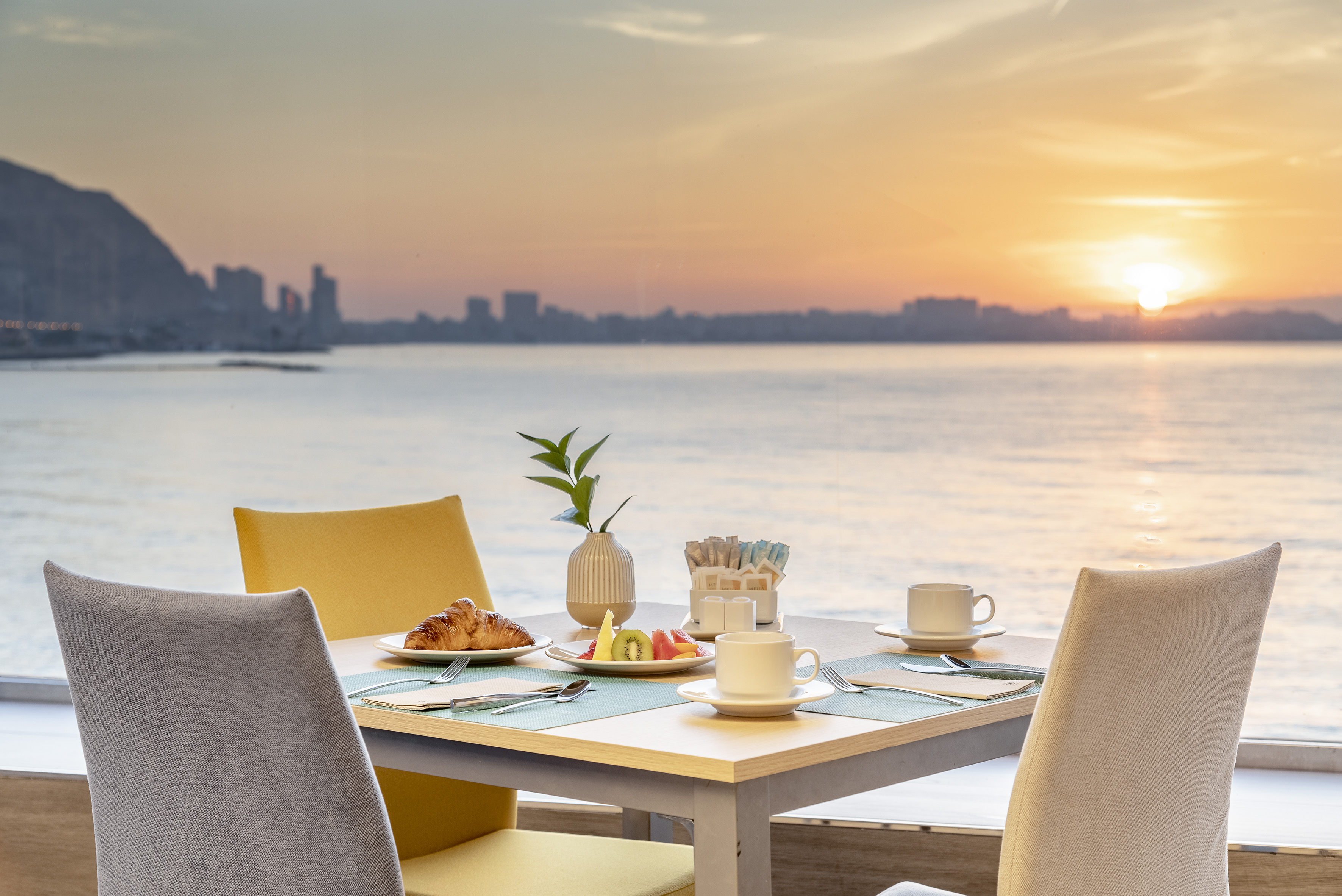 Waterfront breakfast table with croissant, fruit, coffee, and sunrise view.