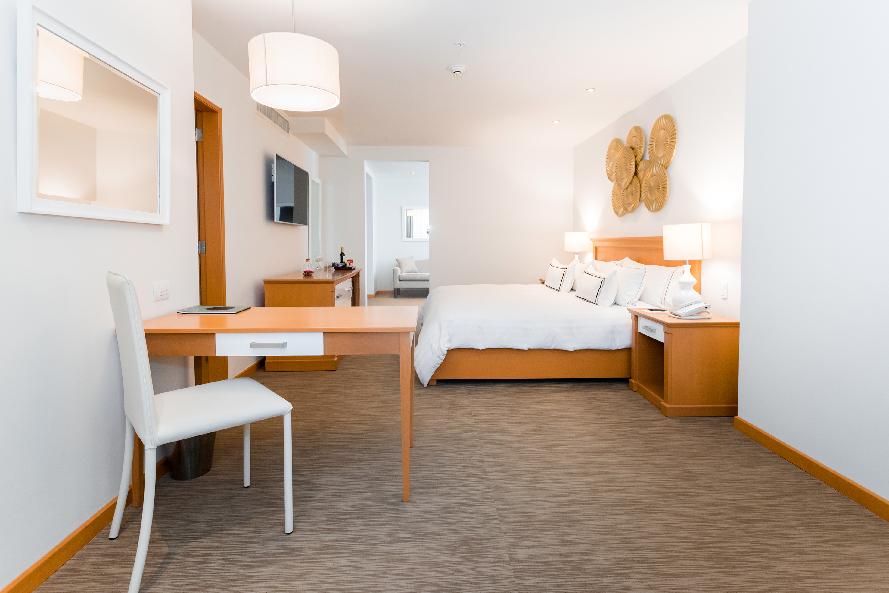 Modern hotel bedroom with white bed, wooden desk, TV, and elegant wall art.