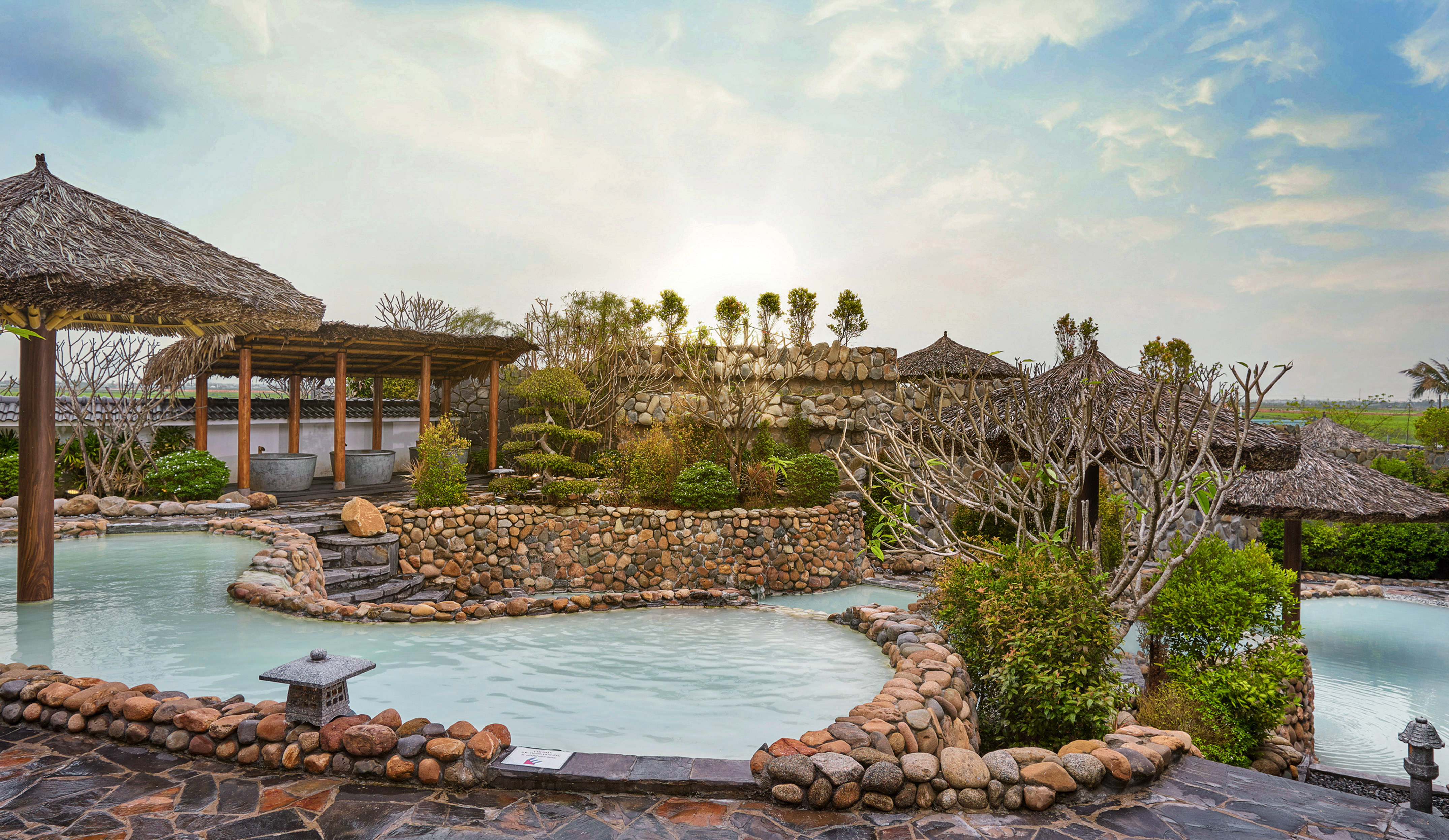 Outdoor hot springs resort with stone pools, thatched roofs, and lush greenery.