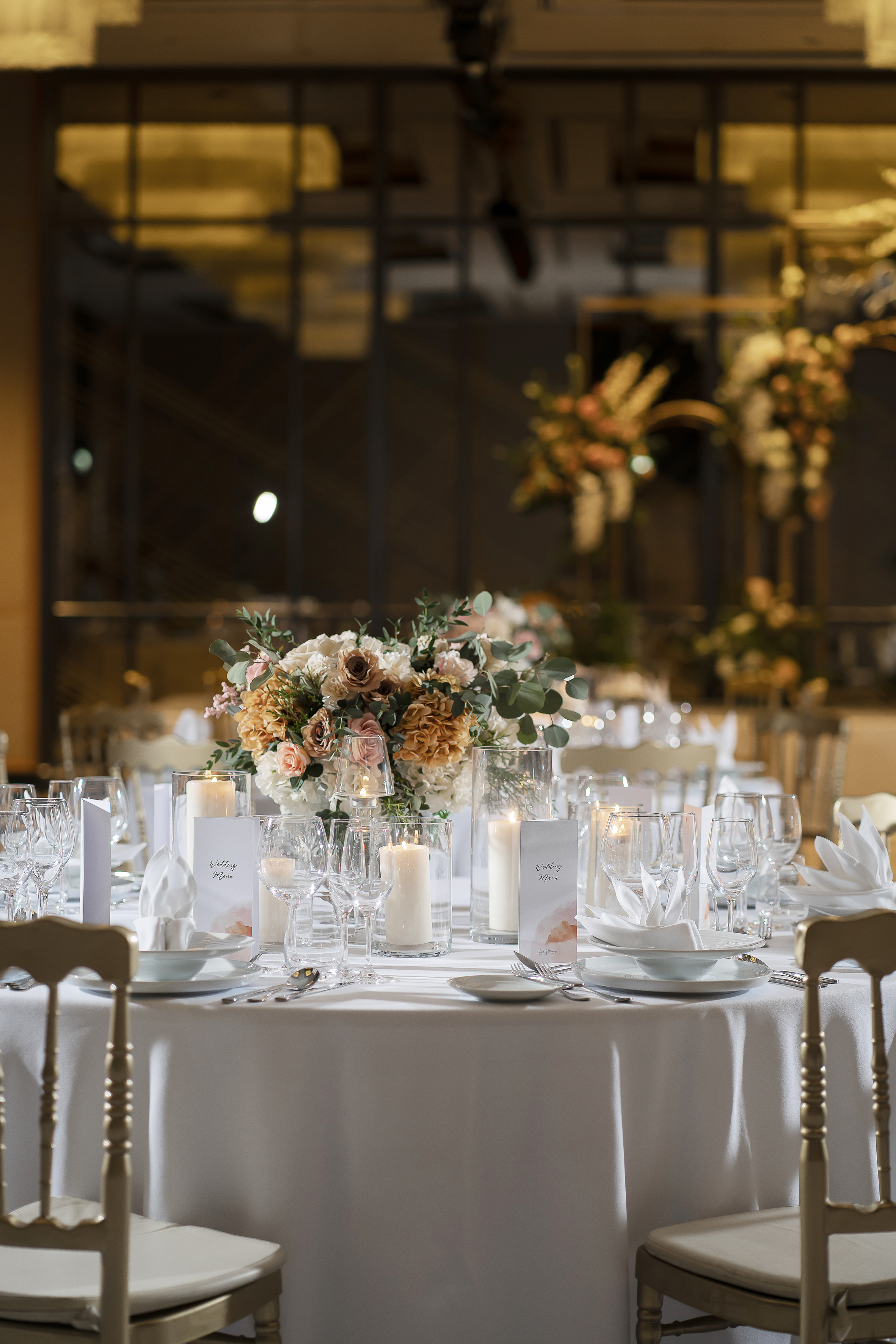 Elegant wedding table with floral centerpiece, candles, menu cards, and gold chairs.