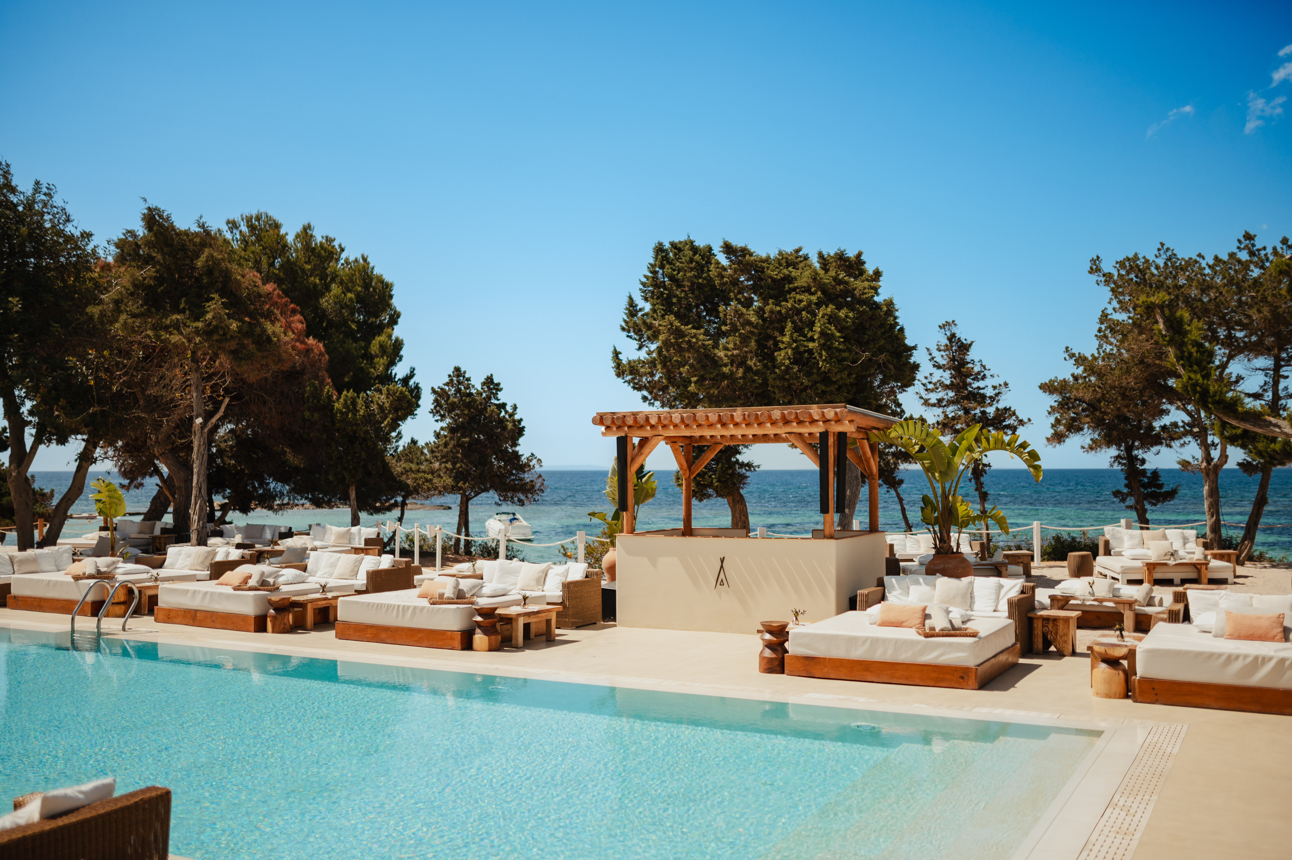 Poolside luxury resort with daybeds, wooden bar, green trees, and blue sea.