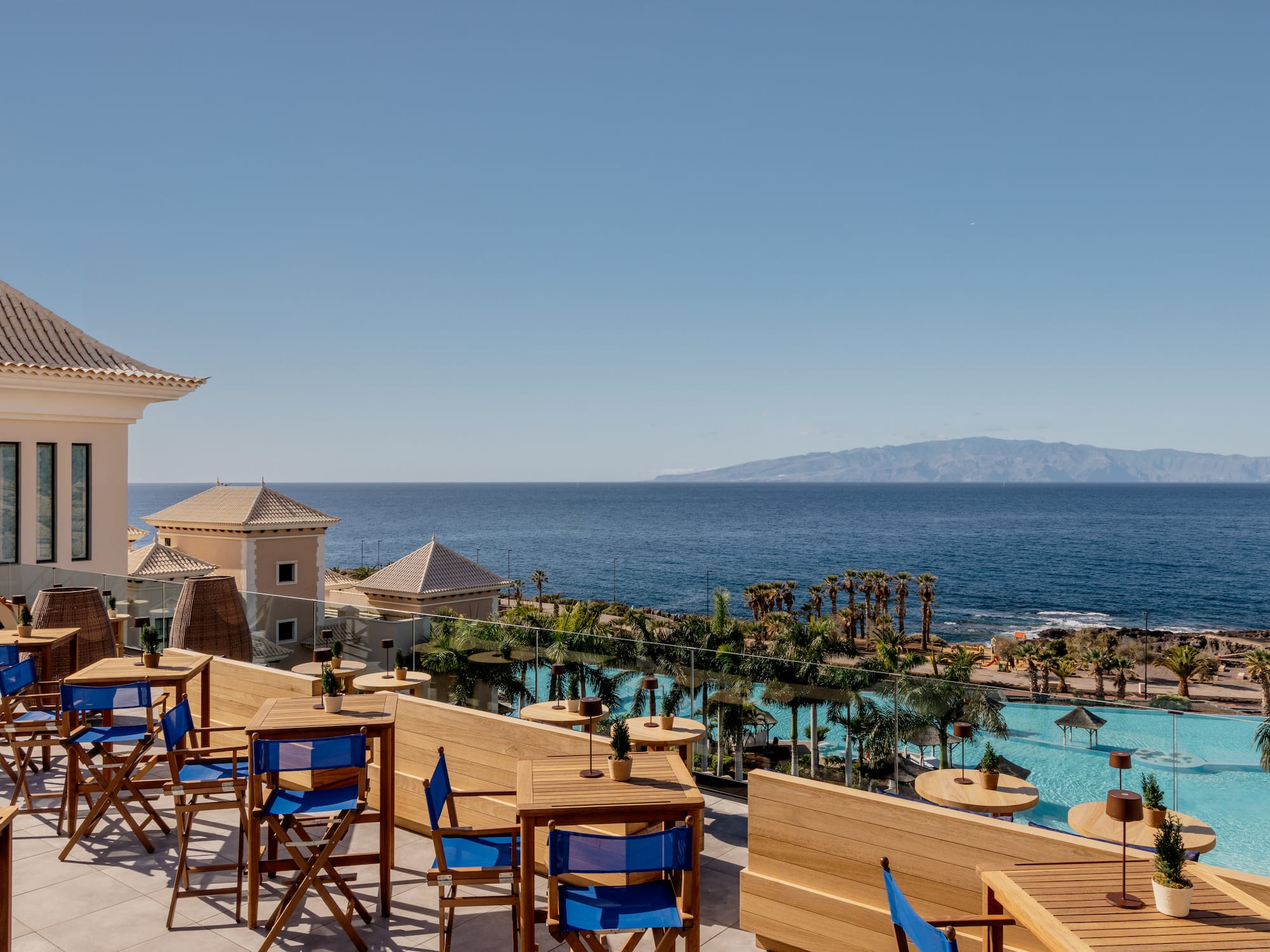 Resort dining terrace overlooking ocean, hotel pool, and distant mountains.