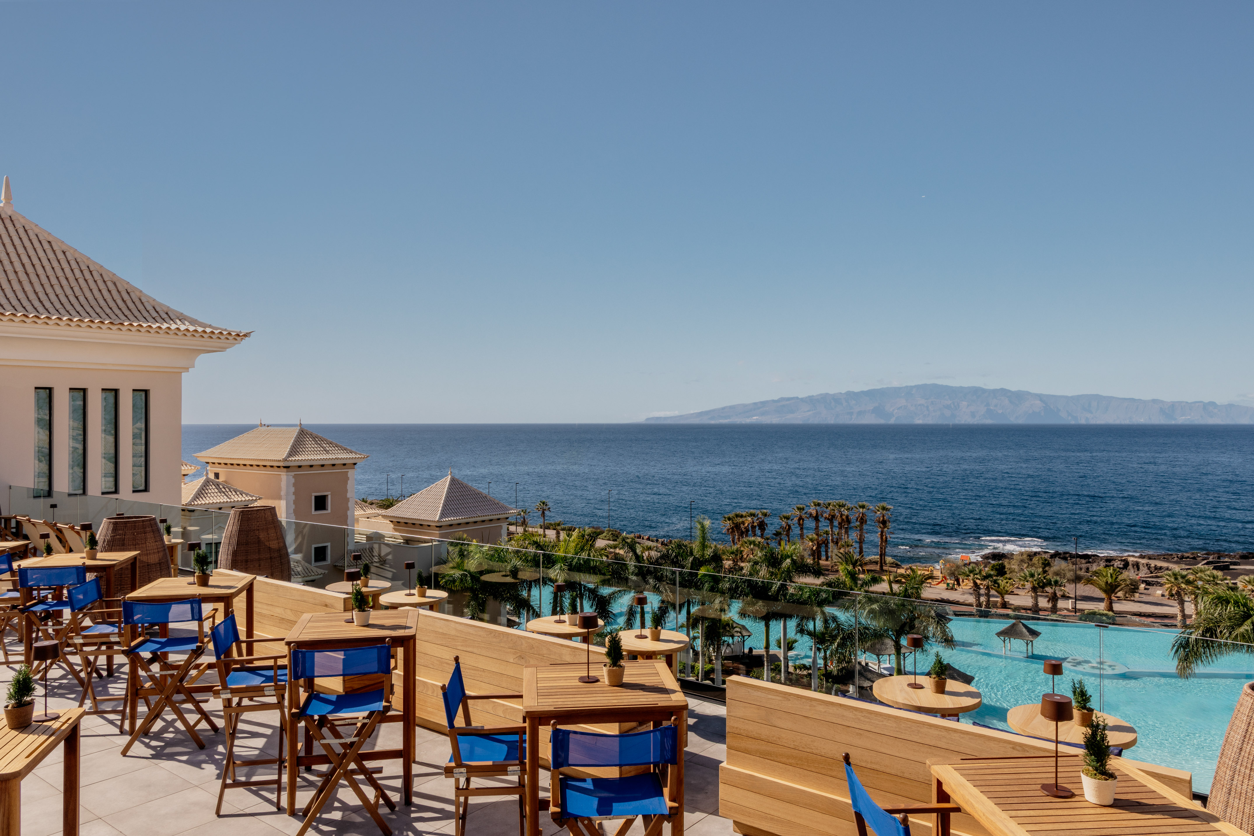 Resort dining terrace overlooking ocean, hotel pool, and distant mountains.