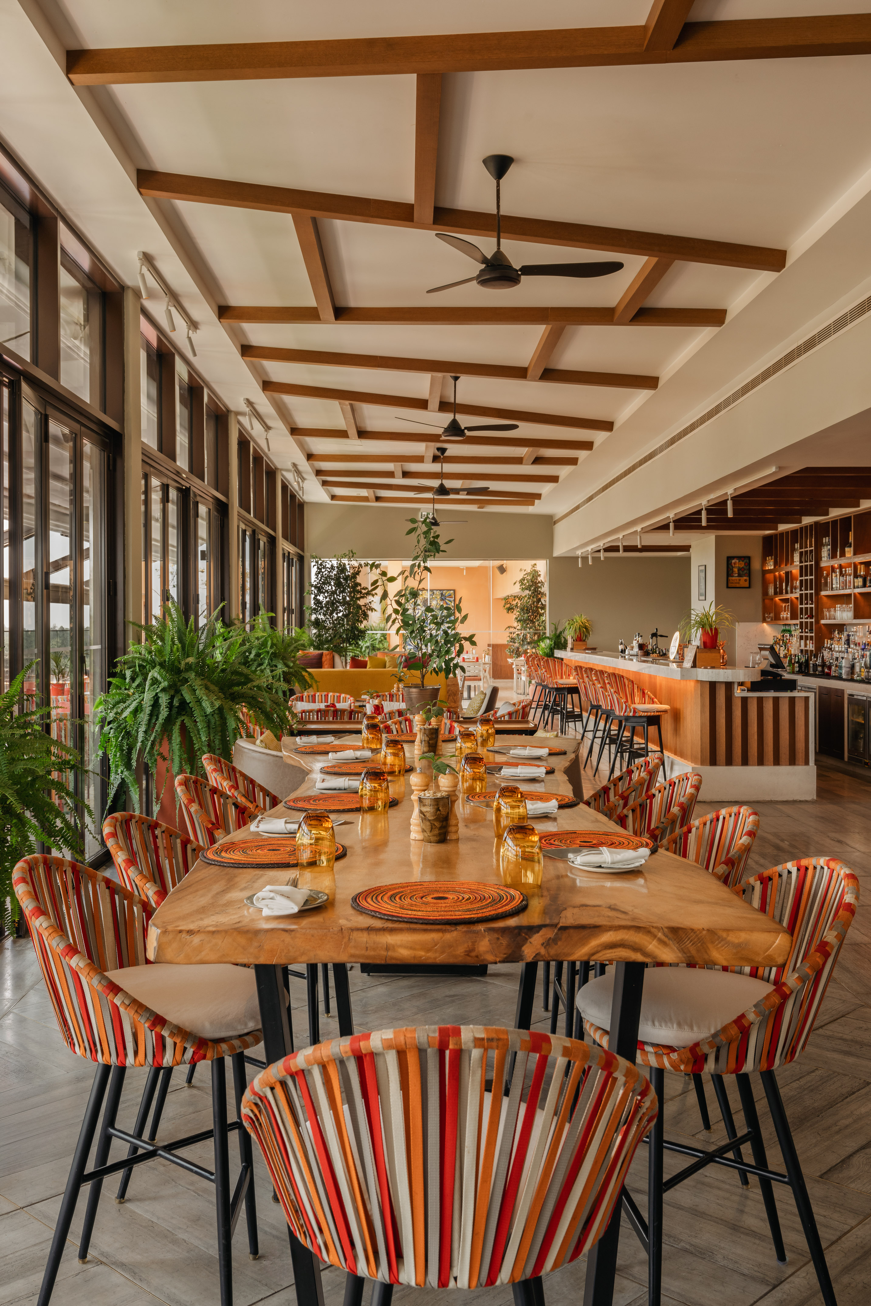 Modern restaurant dining room with wooden table, colorful chairs, and bar.