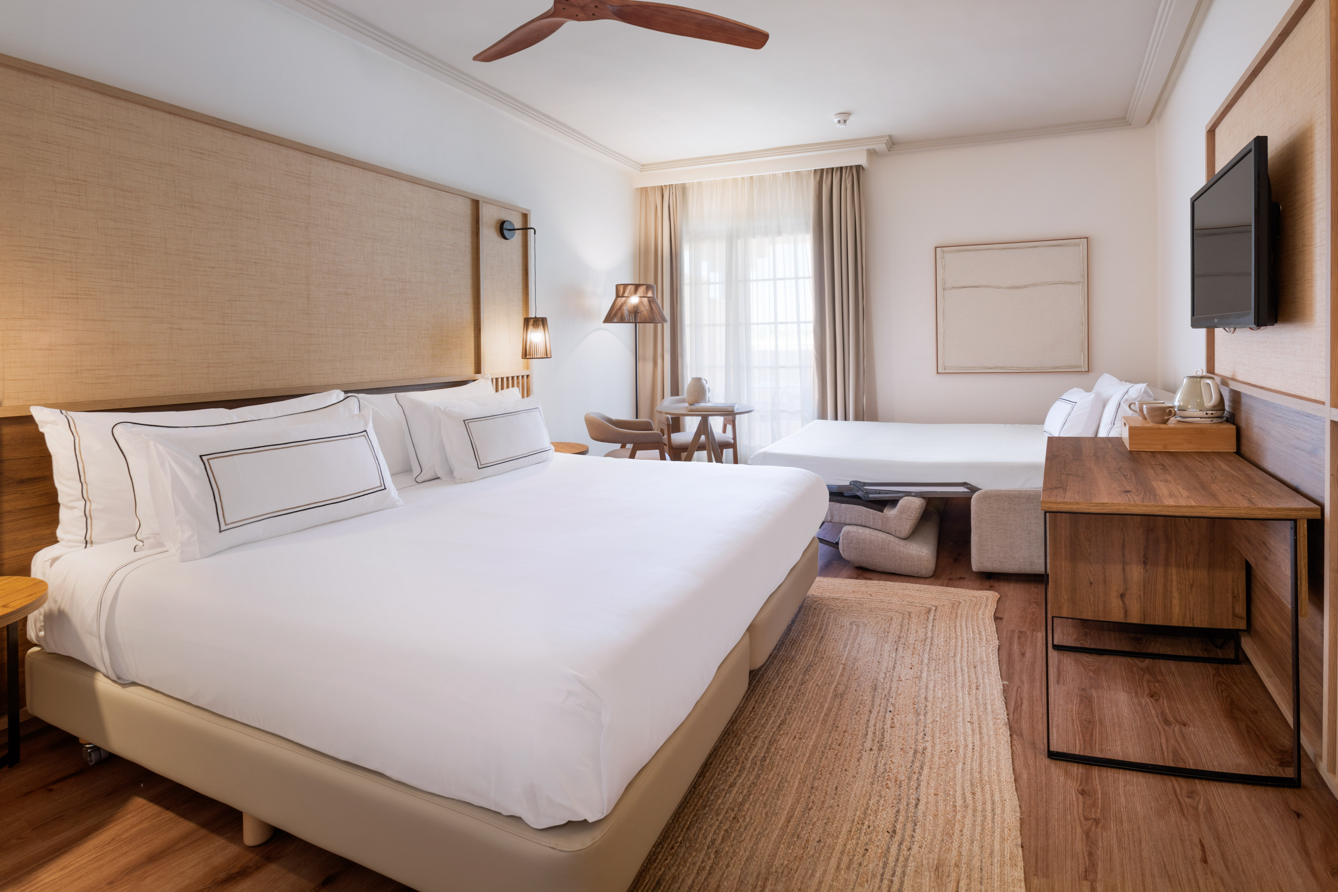 Modern hotel room with king bed, sofa bed, wooden desk, and neutral decor.