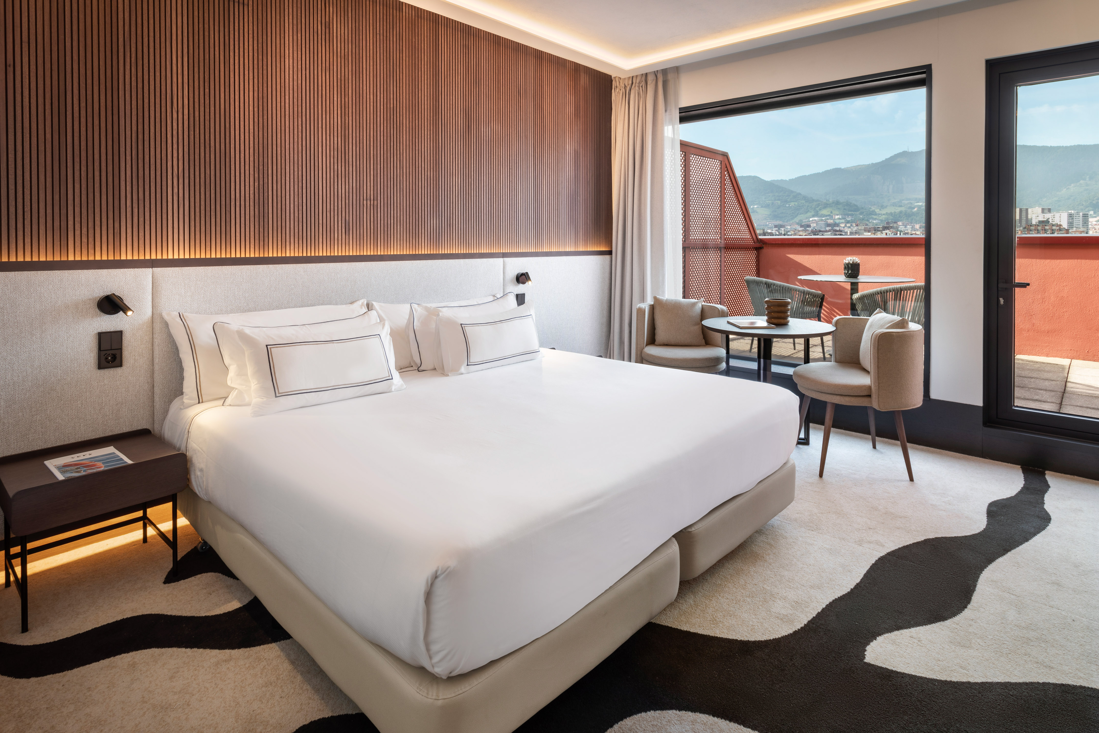 Modern hotel bedroom with white bed, wood wall, balcony, and mountain view.