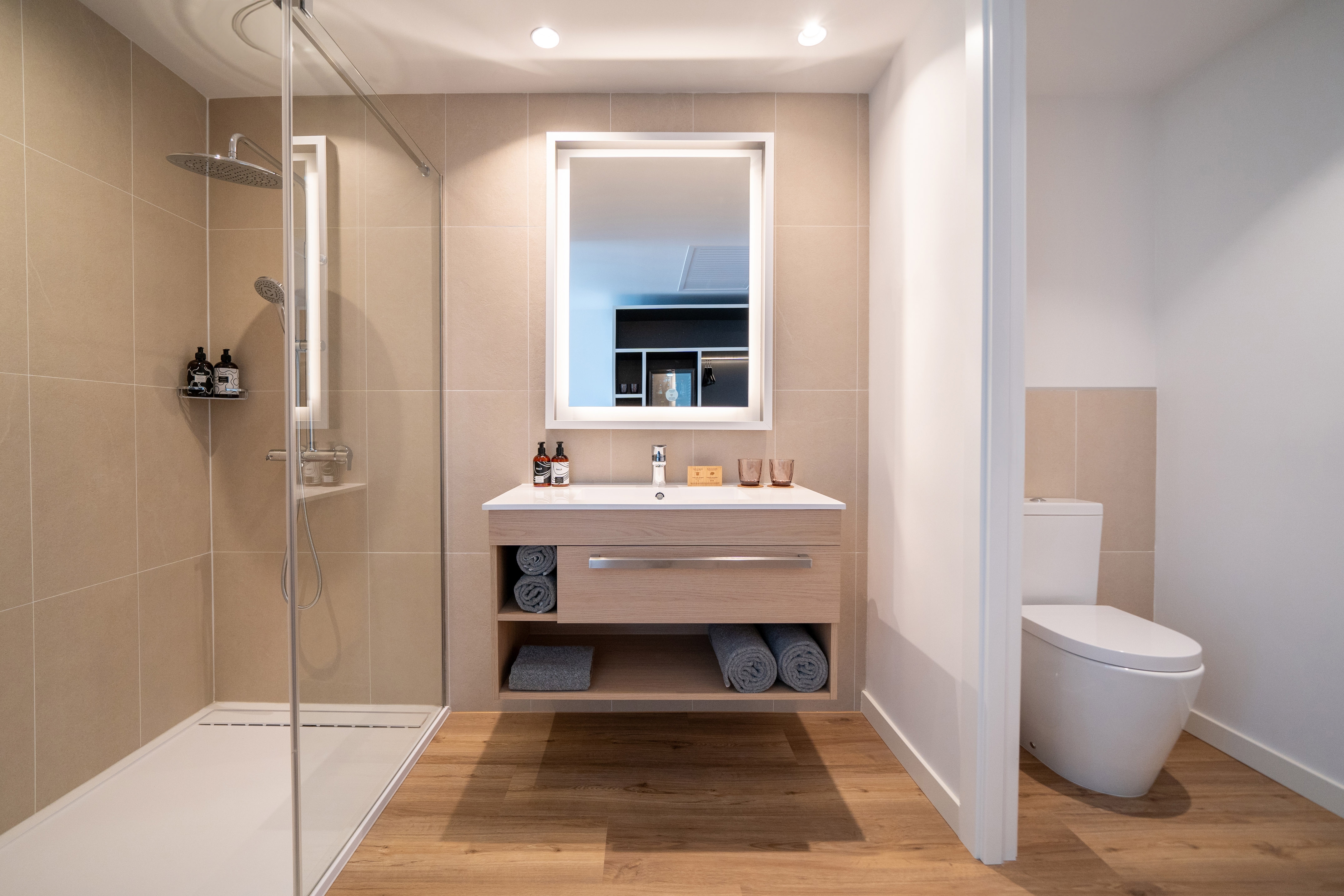 Modern hotel bathroom with walk-in shower, wooden vanity, LED mirror, and toilet.