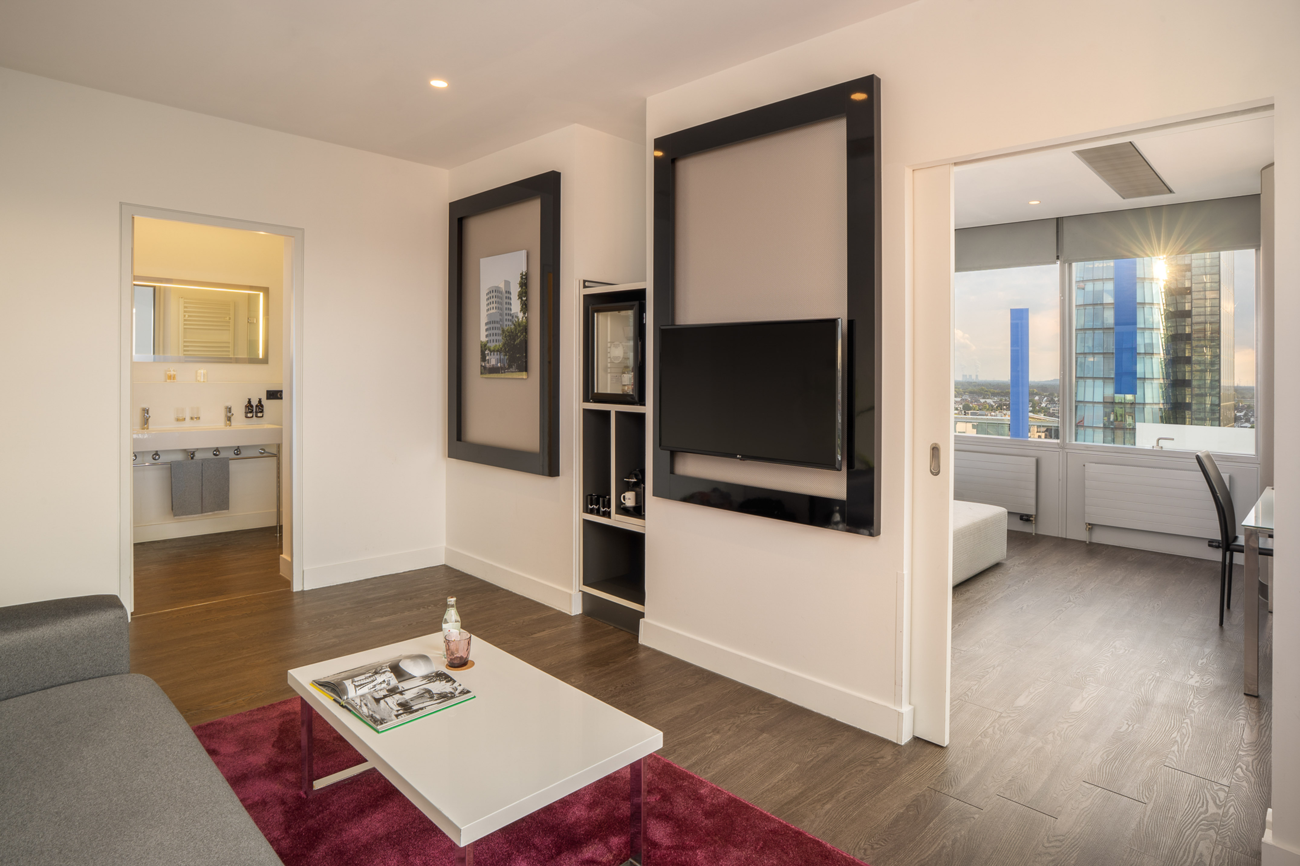 Modern hotel suite living room with city view, sofa, TV, and bathroom.