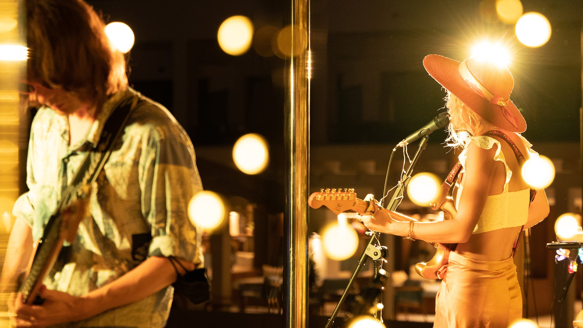 Live performance: Two guitarists on stage, woman in hat with mic, golden bokeh lights.