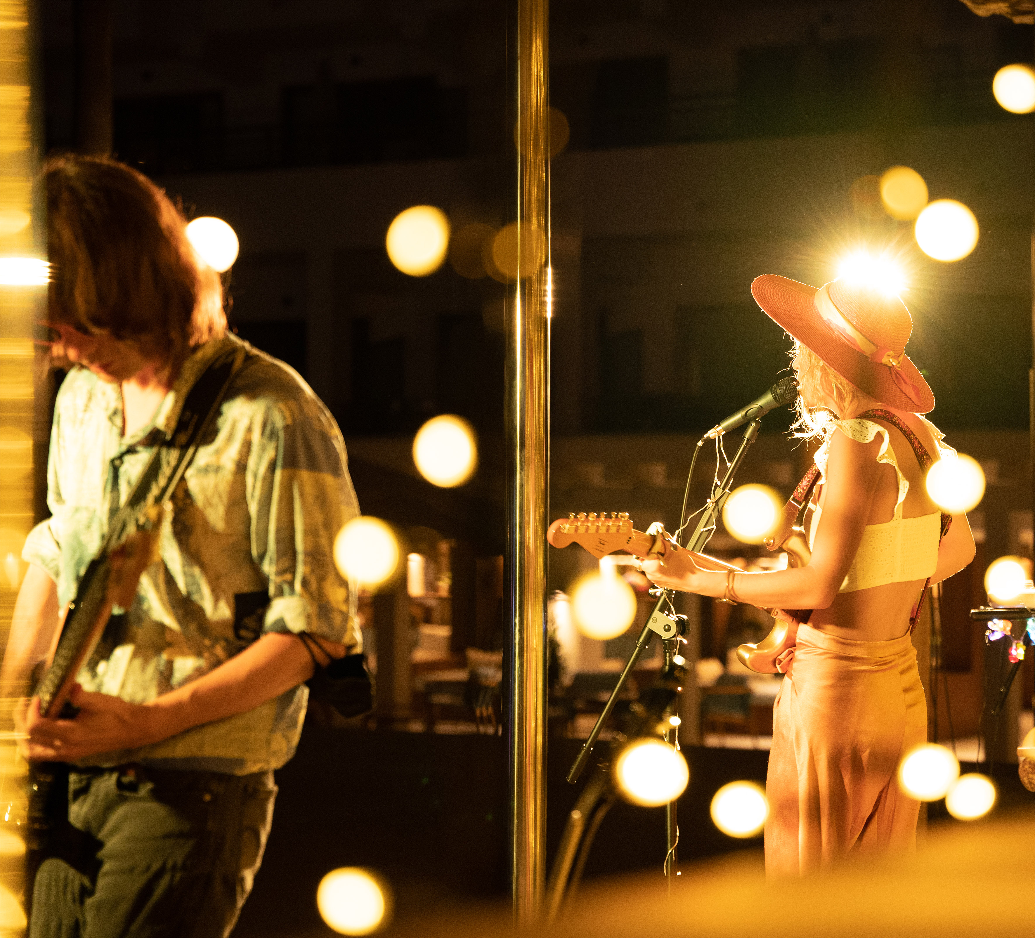 Live performance: Two guitarists on stage, woman in hat with mic, golden bokeh lights.