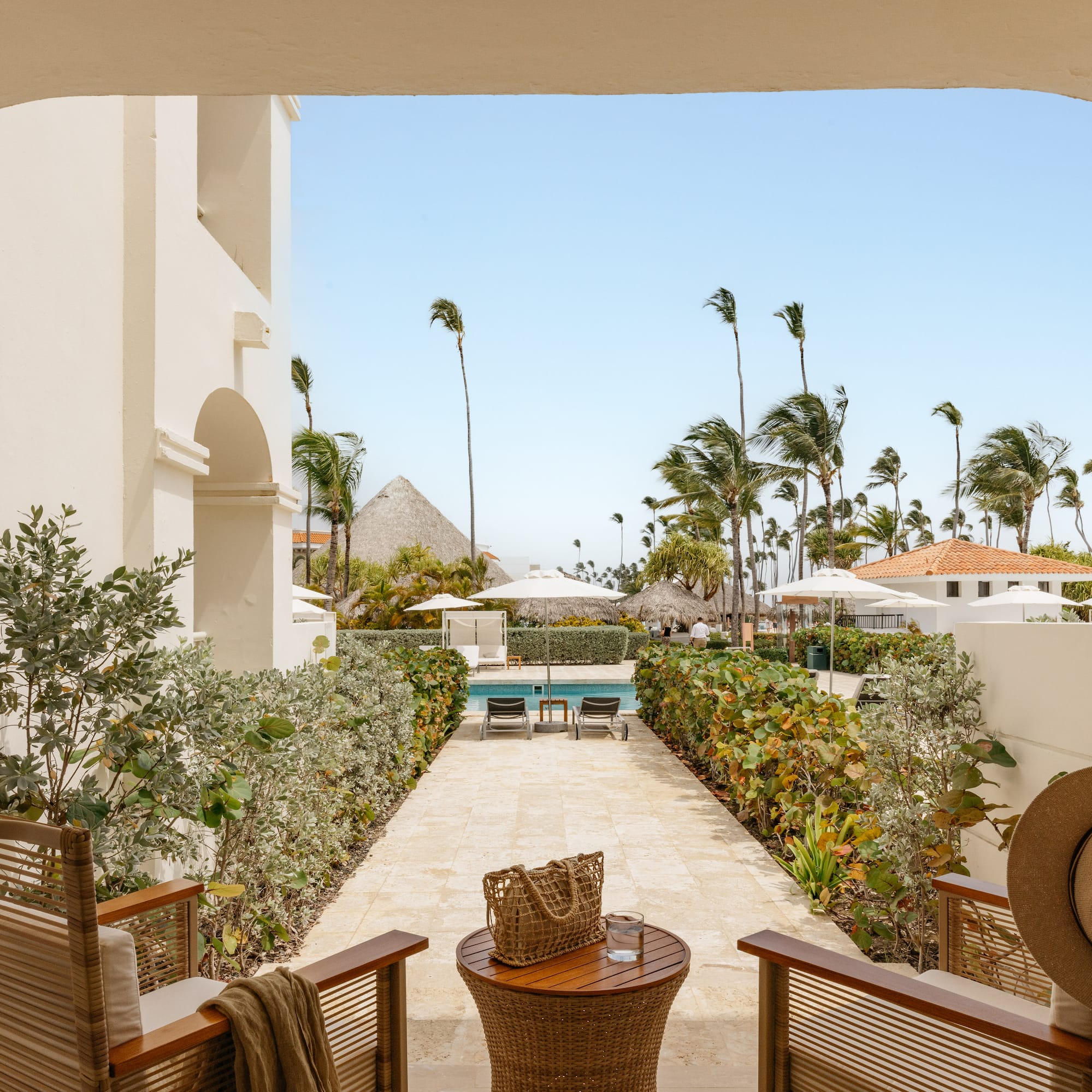 Arched patio overlooking tropical resort pool, swaying palm trees & cabanas.